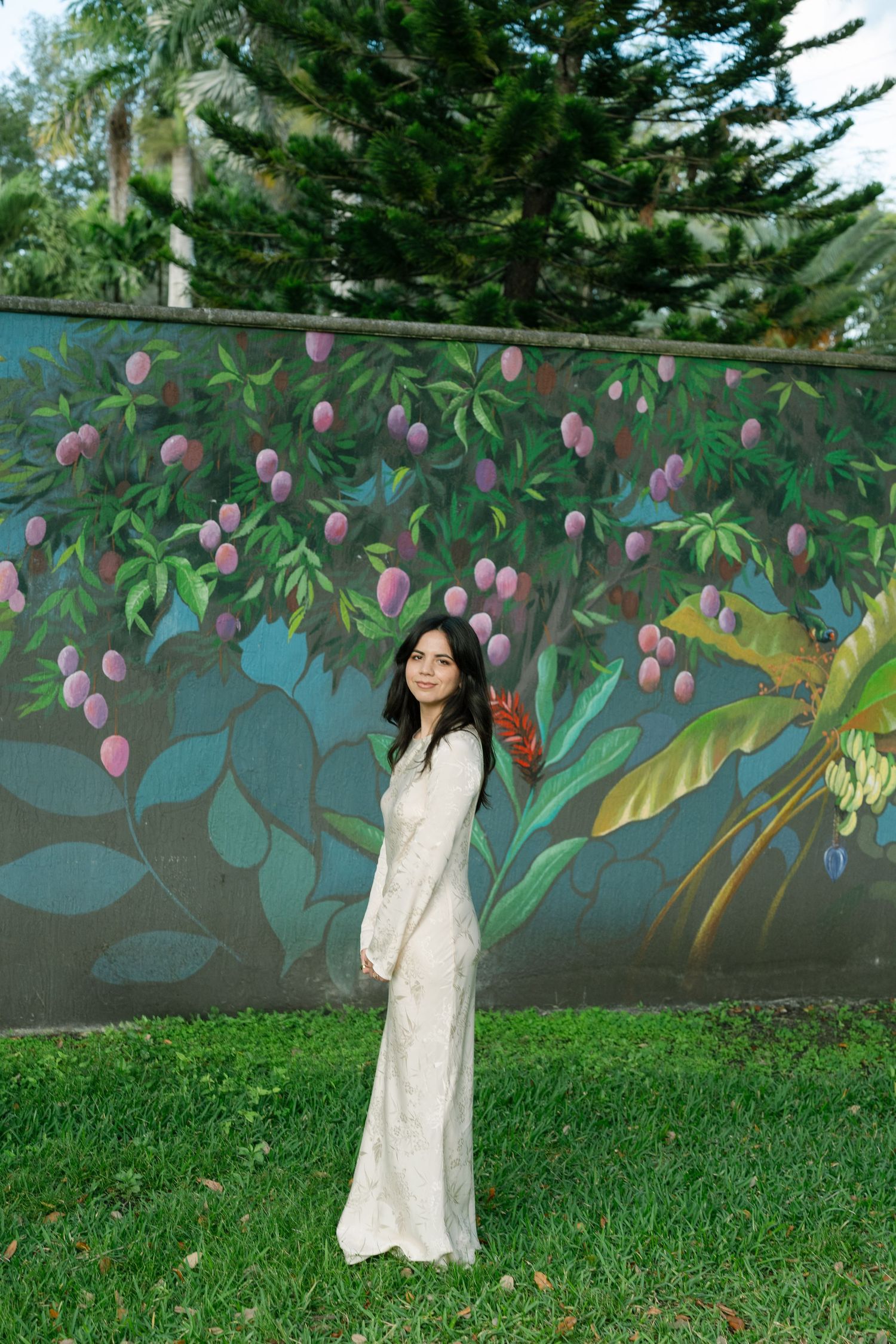 A person in a flowing white dress poses against a colorful mural wall with painted flowers and leaves.