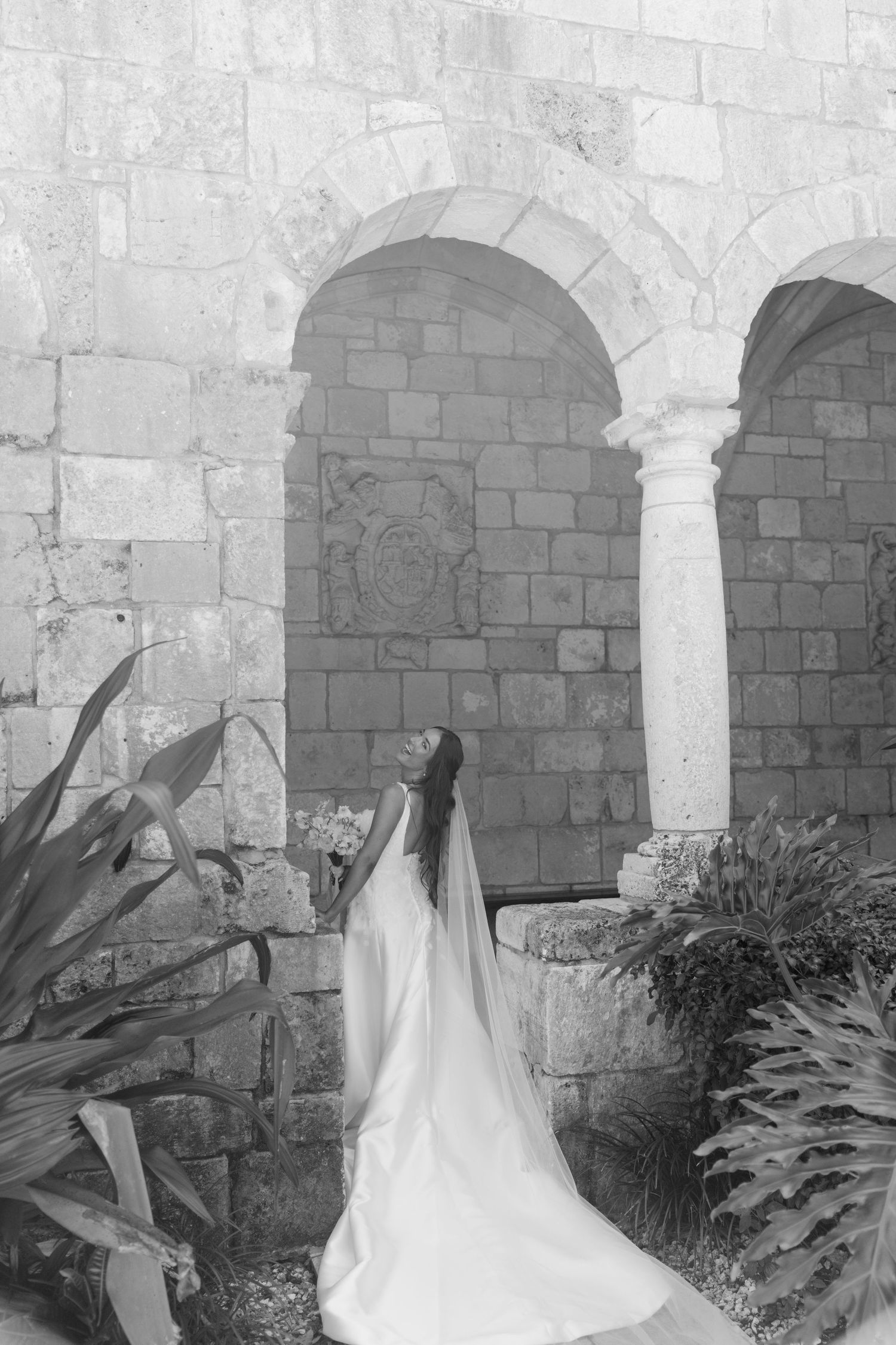 Bride in white gown and veil poses gracefully among stone arches and lush greenery in black and white photo.