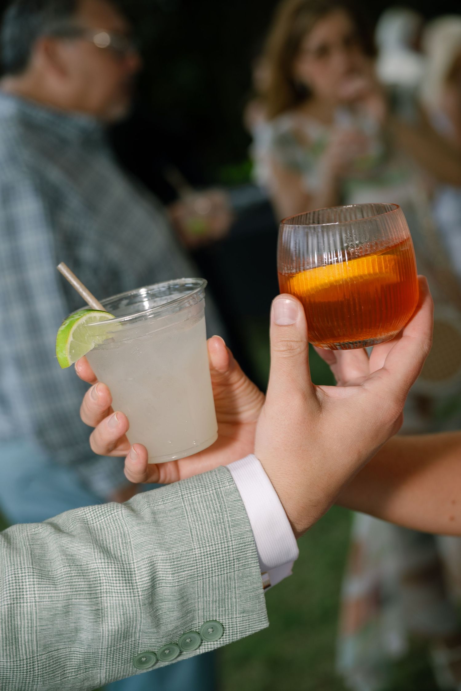 Two hands toasting with cocktails at an outdoor wedding reception gathering.