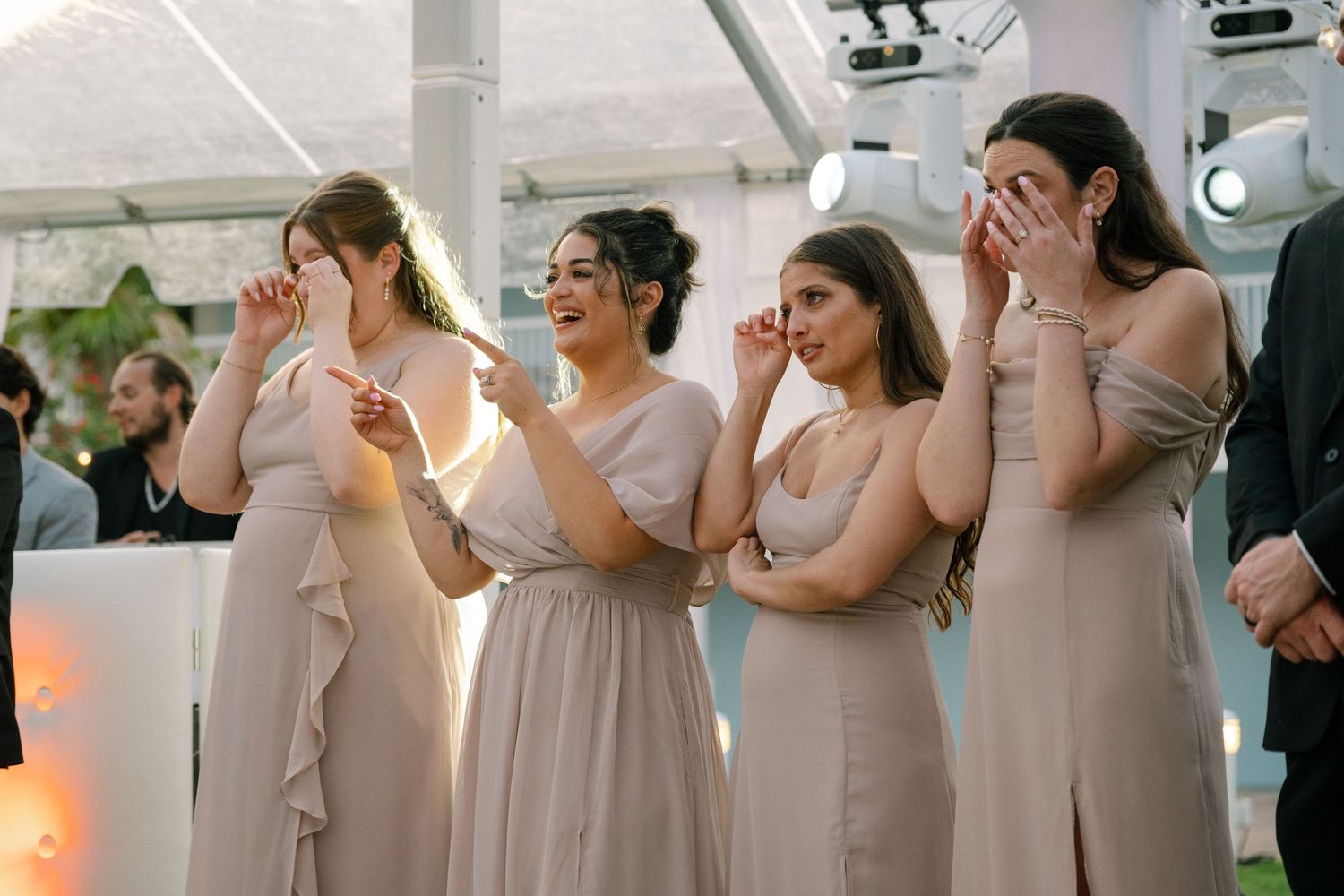 Four emotional bridesmaids in taupe gowns wiping tears during a heartfelt wedding reception moment under a tent.