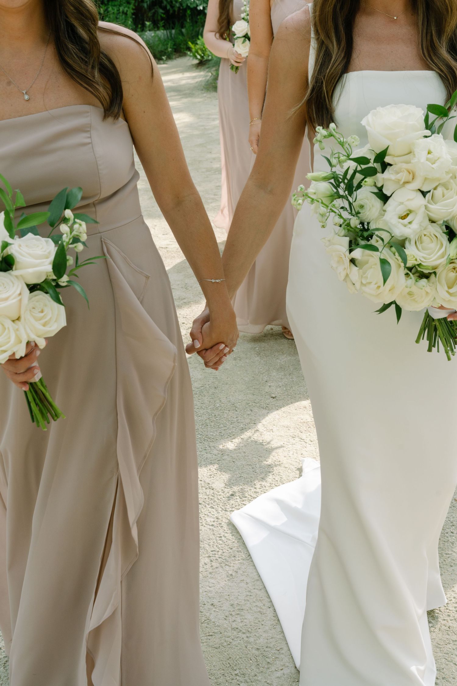 Bride in white gown holding hands with bridesmaid in taupe dress, both carrying white rose bouquets.
