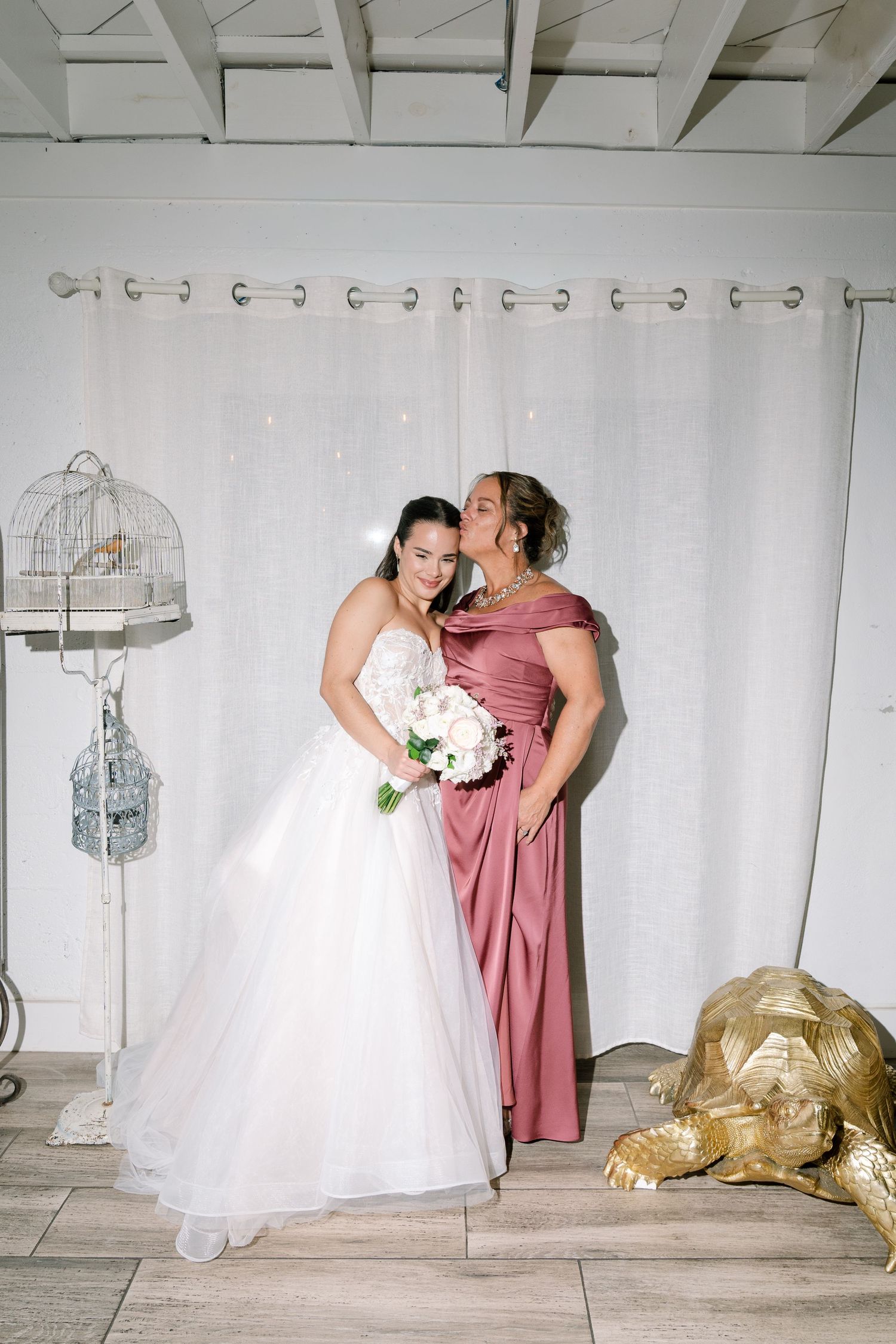 Bride in white ball gown receives kiss on cheek from woman in dusty rose dress at wedding reception.
