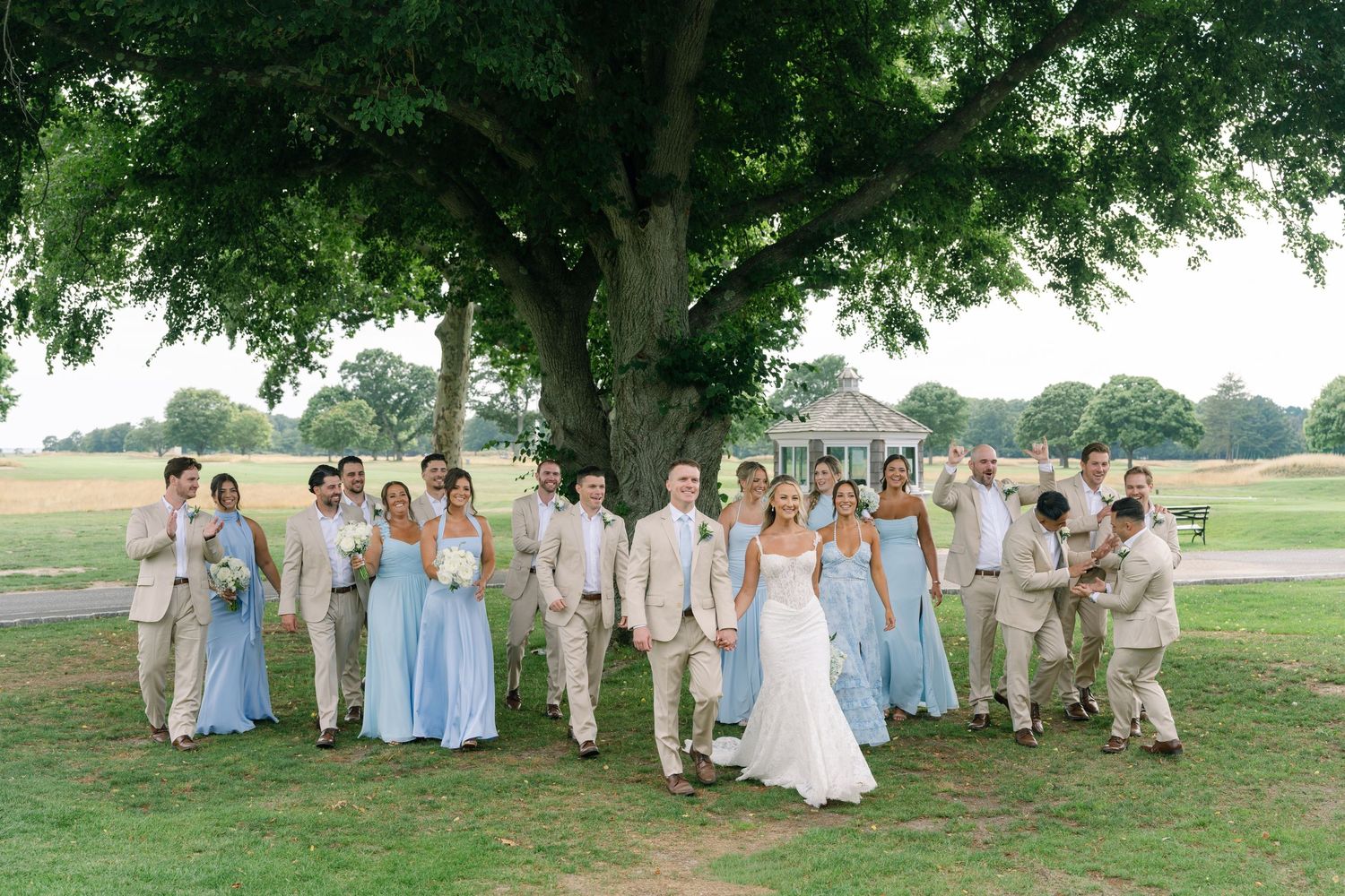 Large wedding party poses under a grand tree, bridesmaids in blue gowns and groomsmen in tan suits.