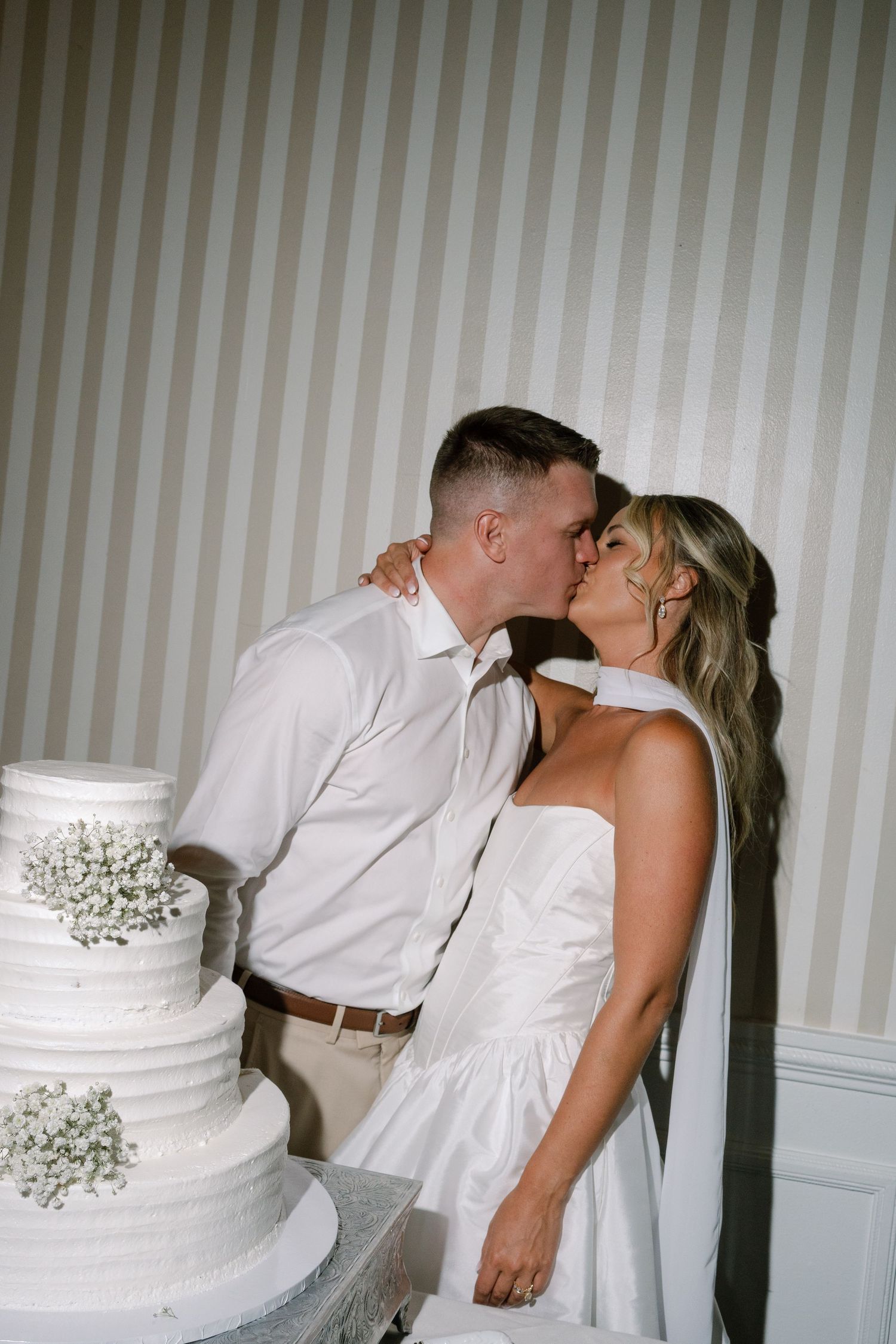 Bride and groom kissing beside elegant white four-tier wedding cake with baby's breath decorations.