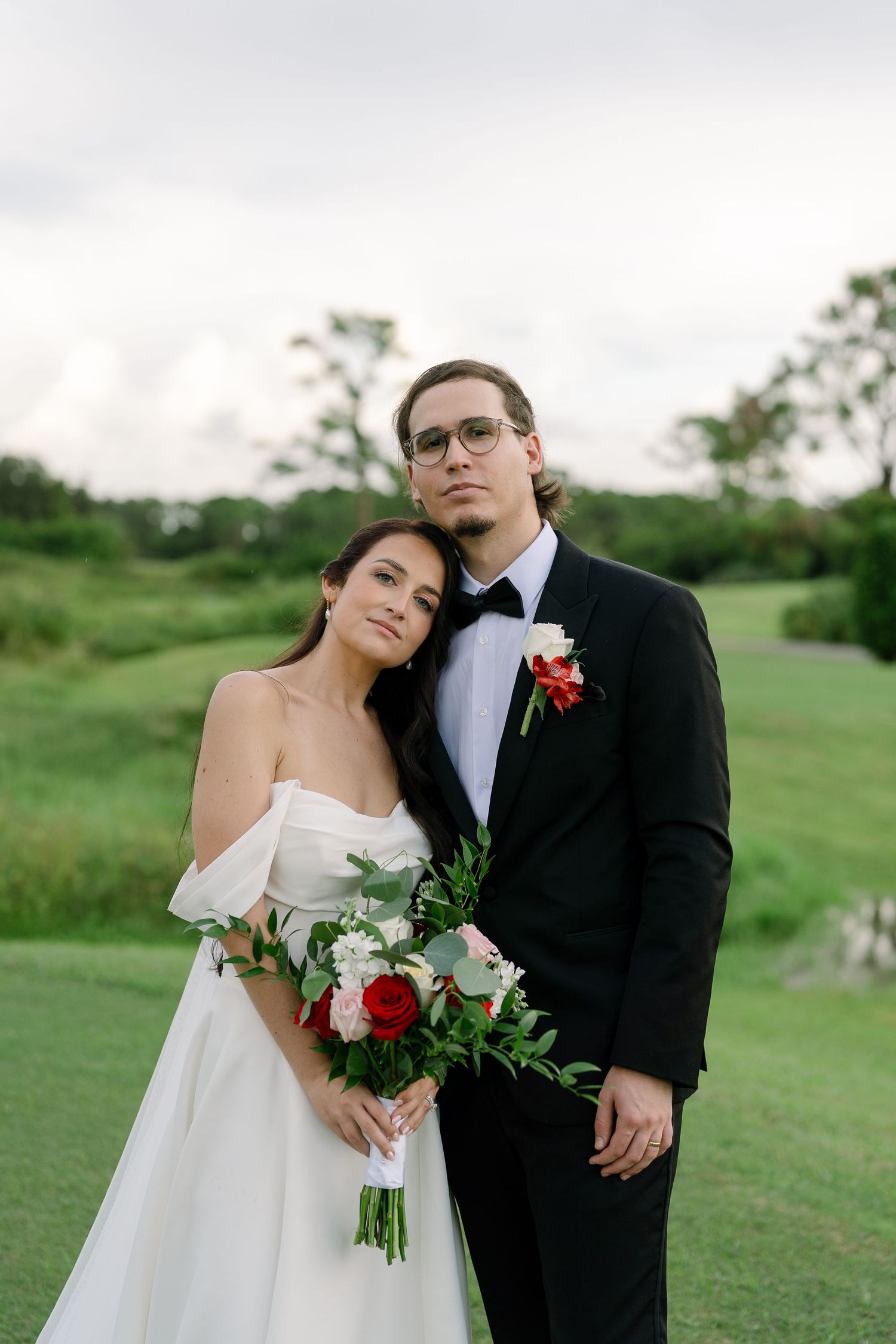 Bride in white off-shoulder gown holds red bouquet beside groom in black tuxedo on lush green golf course.