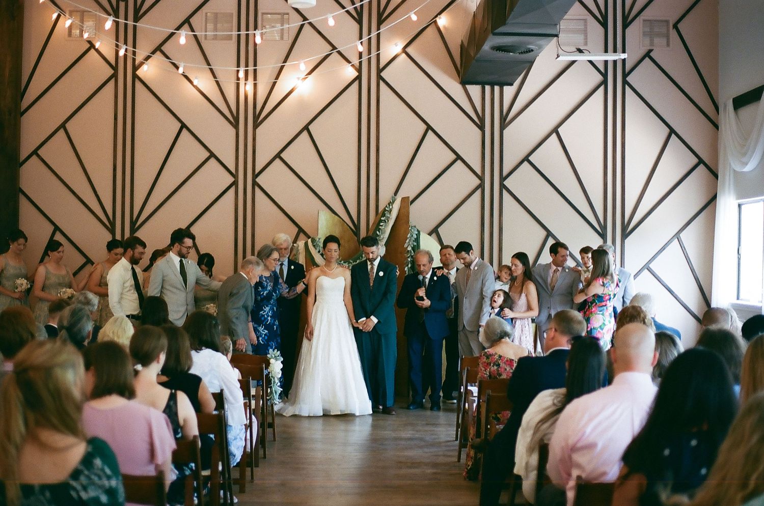 Wedding guests gather in a rustic-style venue with dramatic geometric wooden beams and warm lighting during a reception.