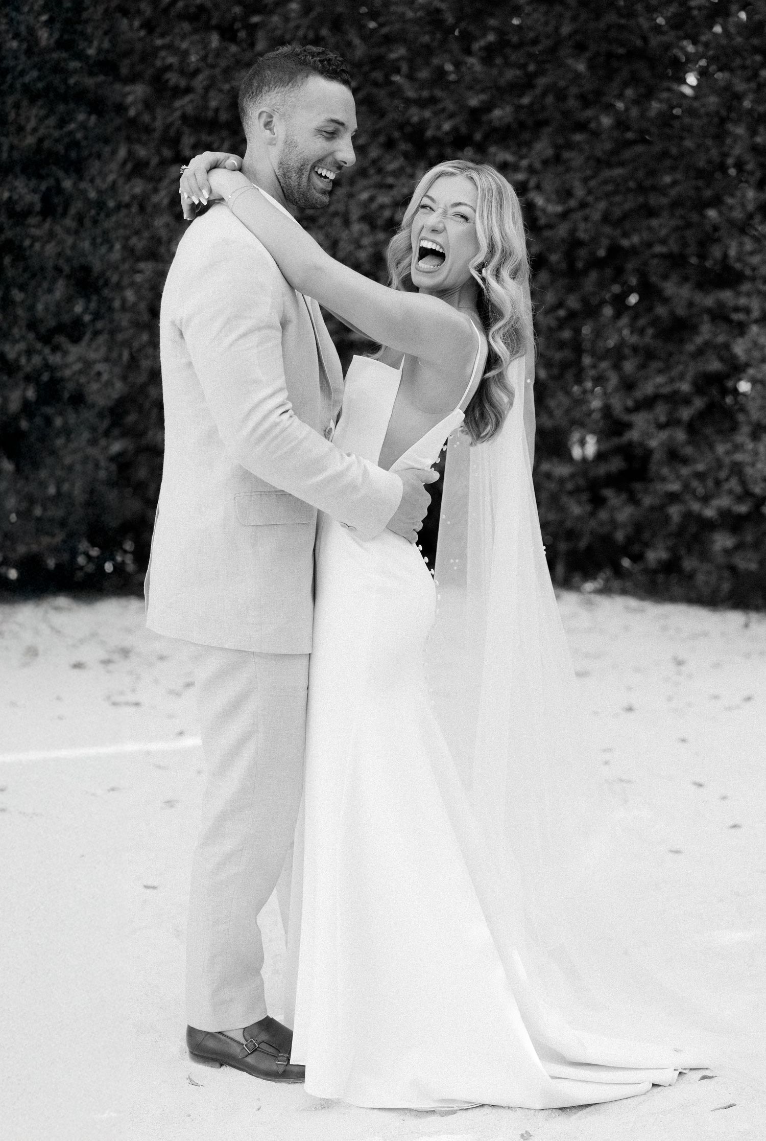 Black and white photo series of a joyful couple in wedding attire dancing and laughing together on a beach.