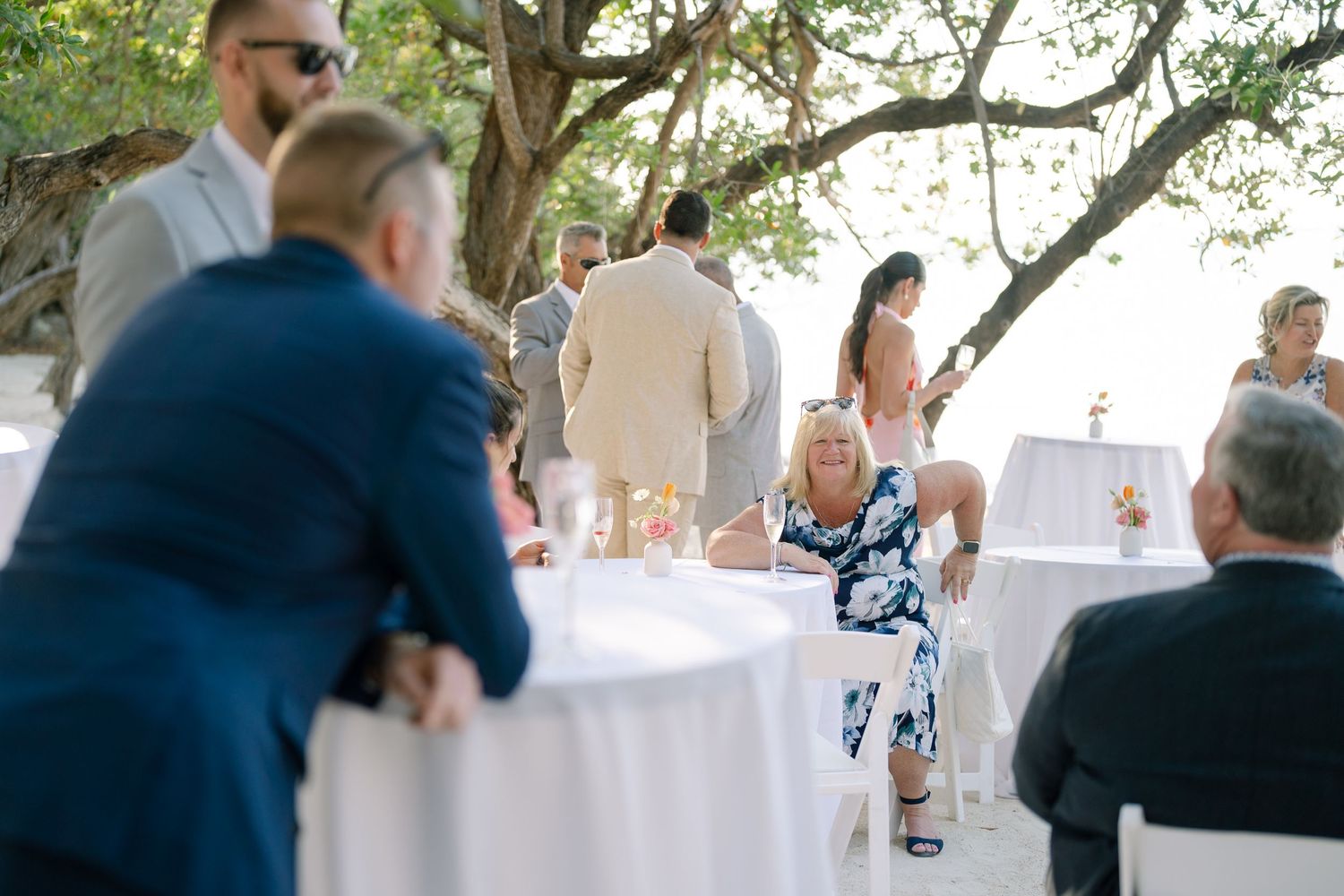 Outdoor wedding reception with guests mingling under trees and white table settings.