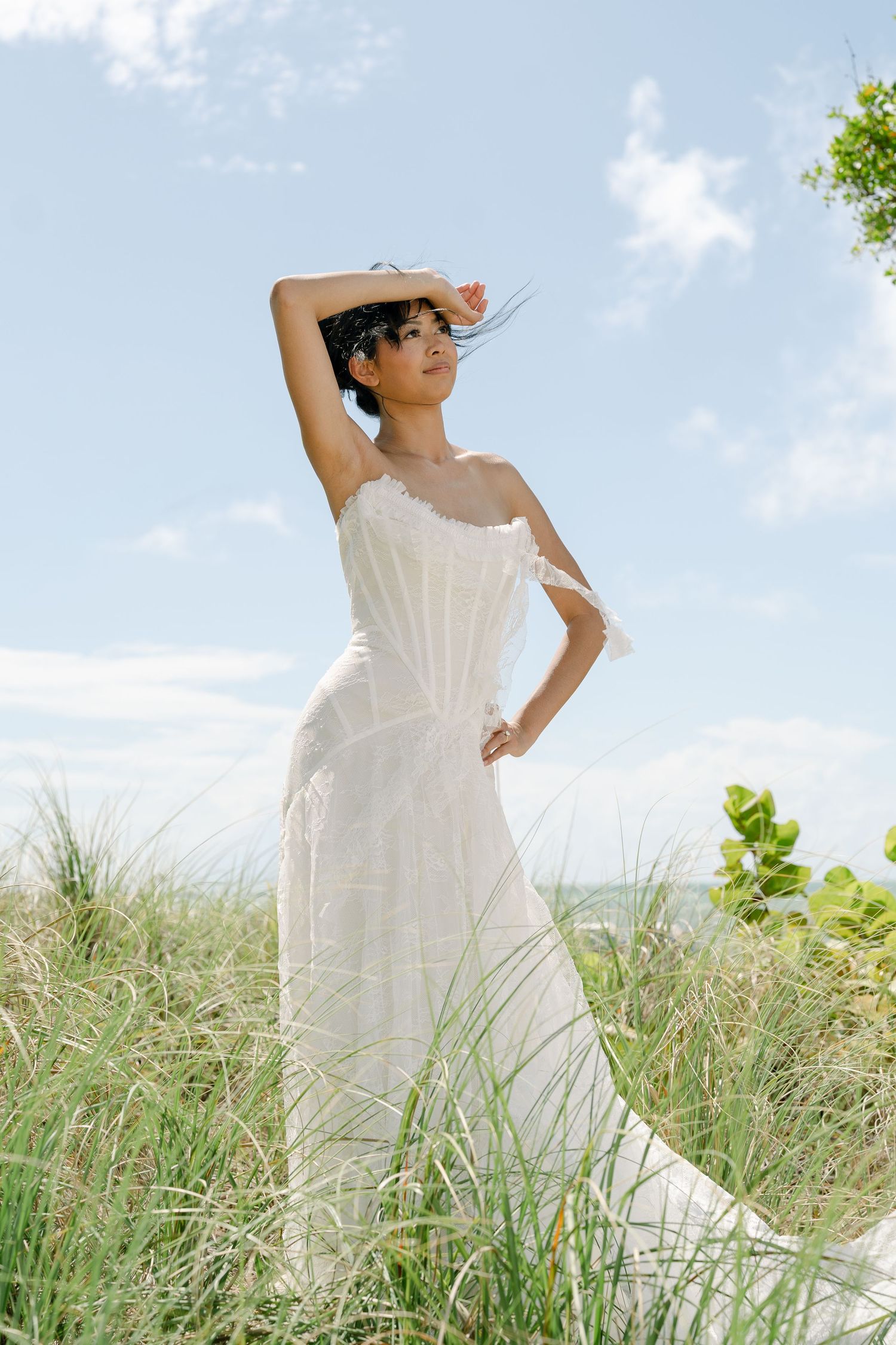 Woman in white strapless dress posing gracefully in coastal grass dunes under a bright blue sky.