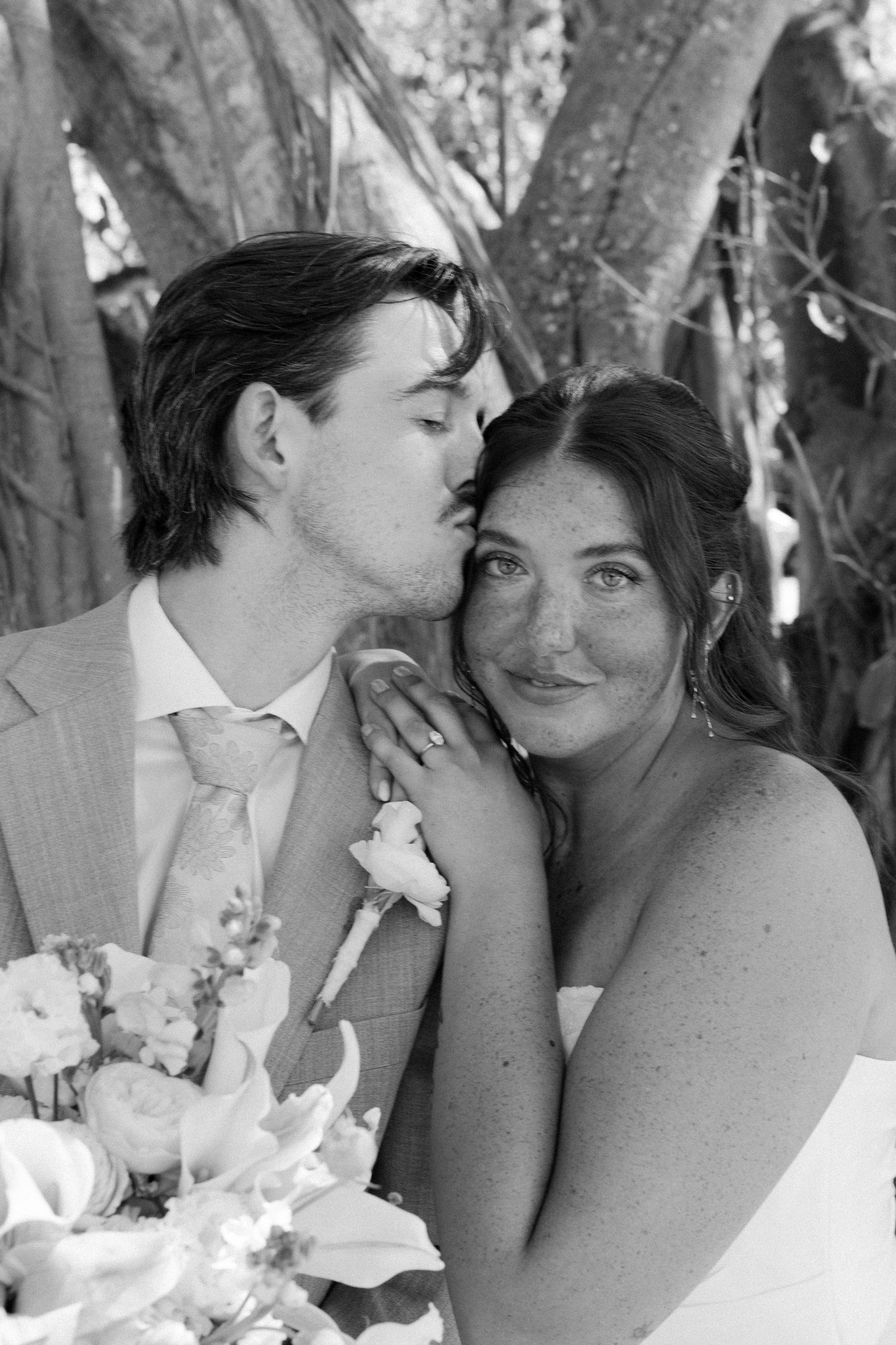 A bride and groom embracing and kissing in an outdoor setting with trees and foliage.