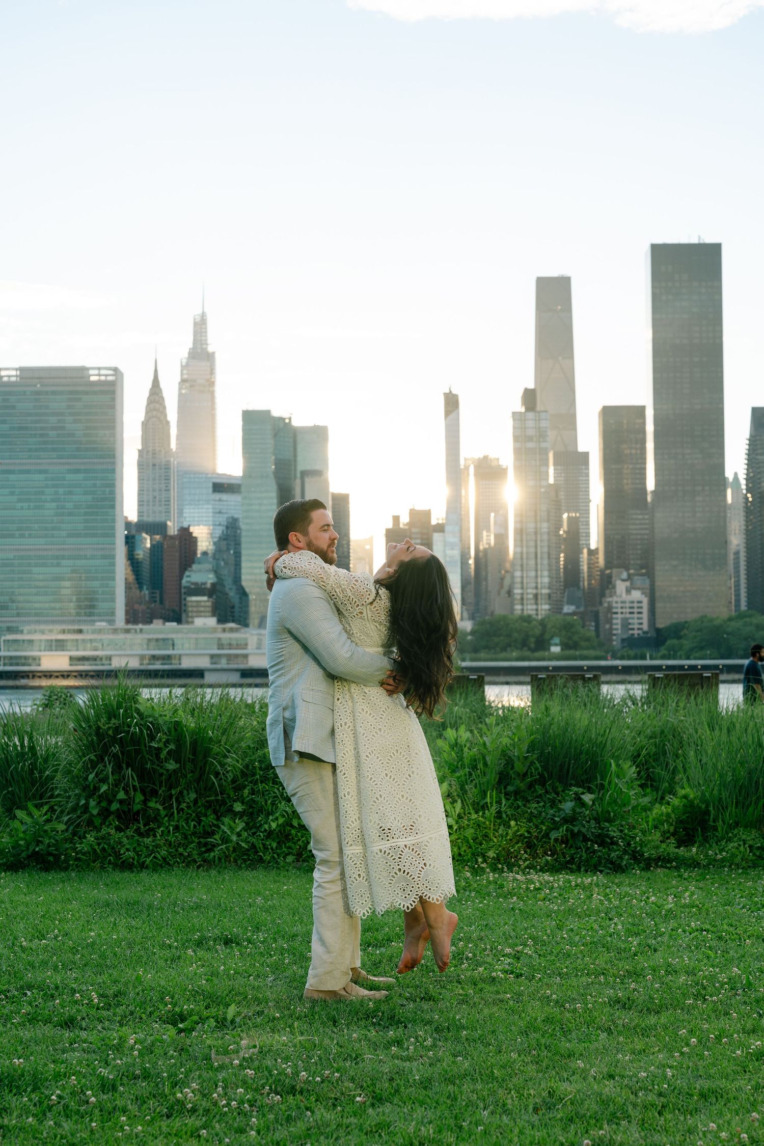 Couple embracing on green lawn with Manhattan skyline and golden sunset behind them in New York City.
