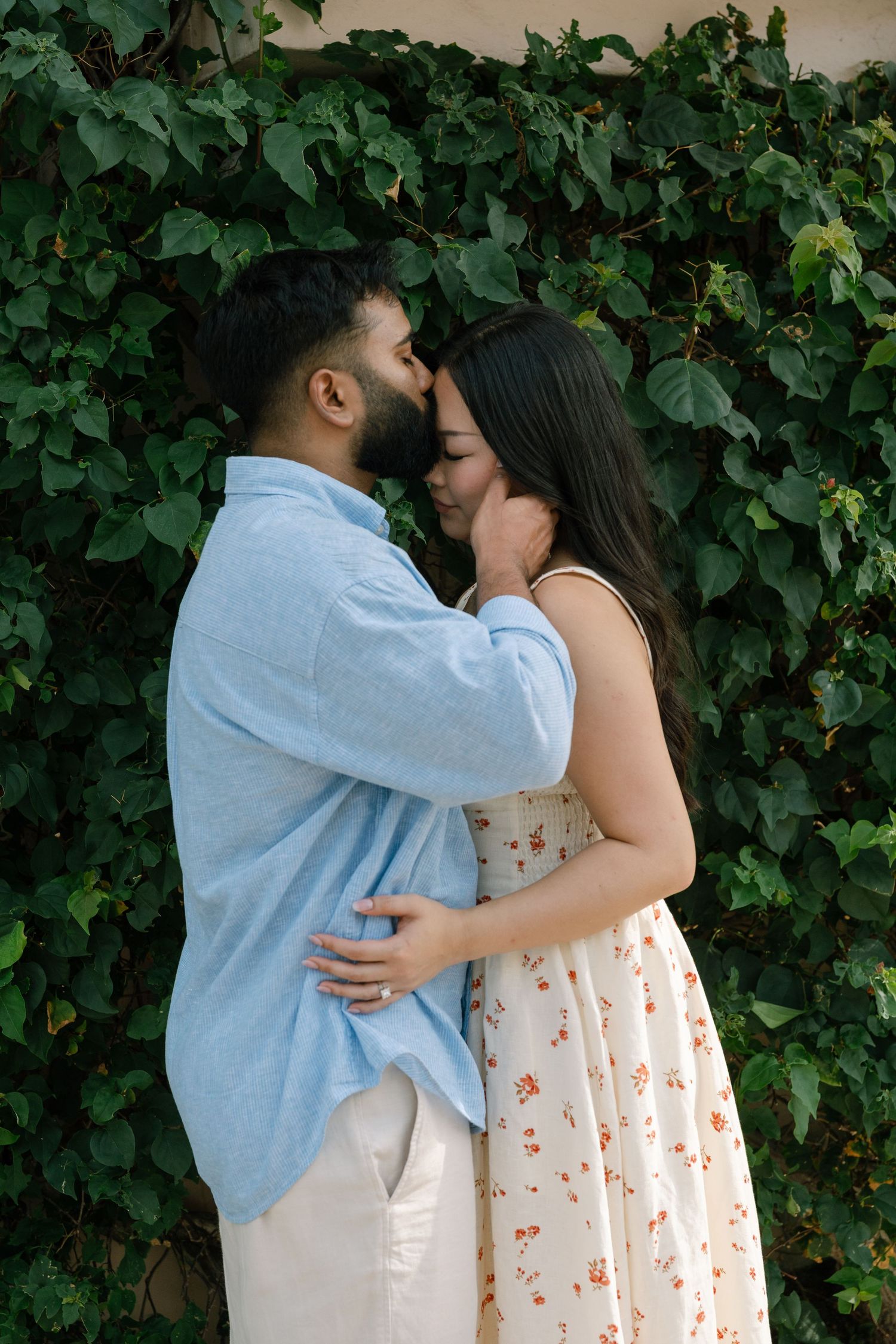 A couple shares a tender moment, embracing and touching foreheads before a lush green ivy-covered wall outdoors.