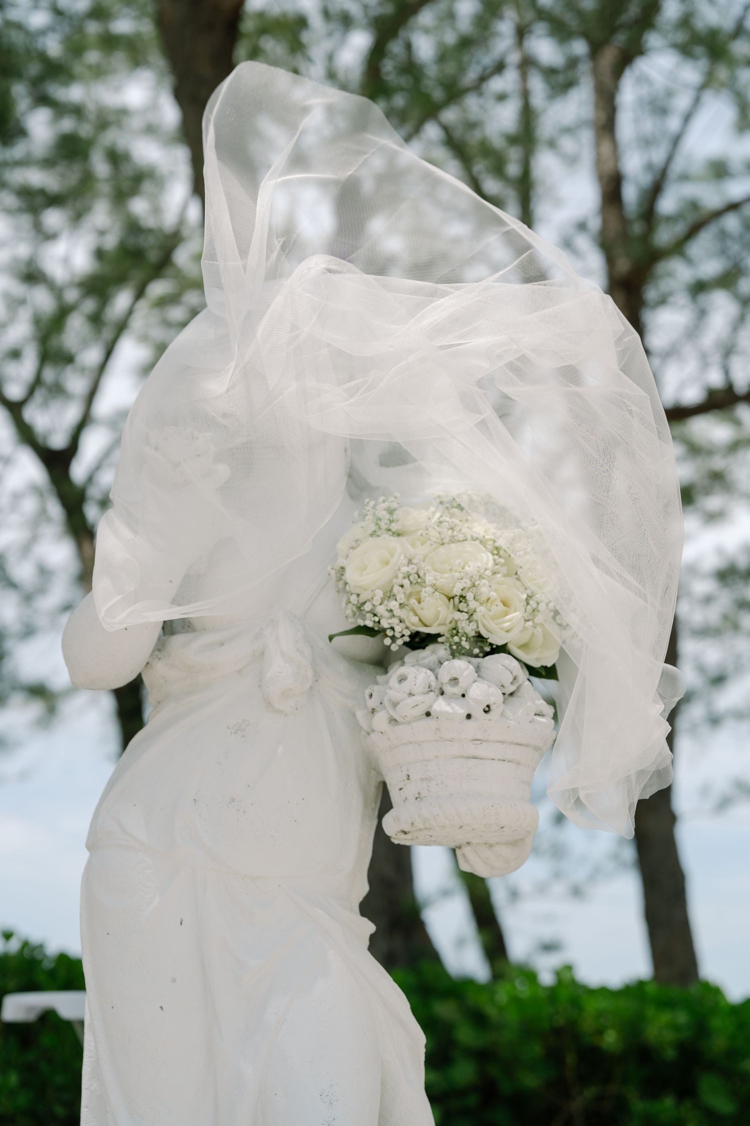 Bride in white wedding dress with veil blowing in wind holding bouquet of white roses outdoors.