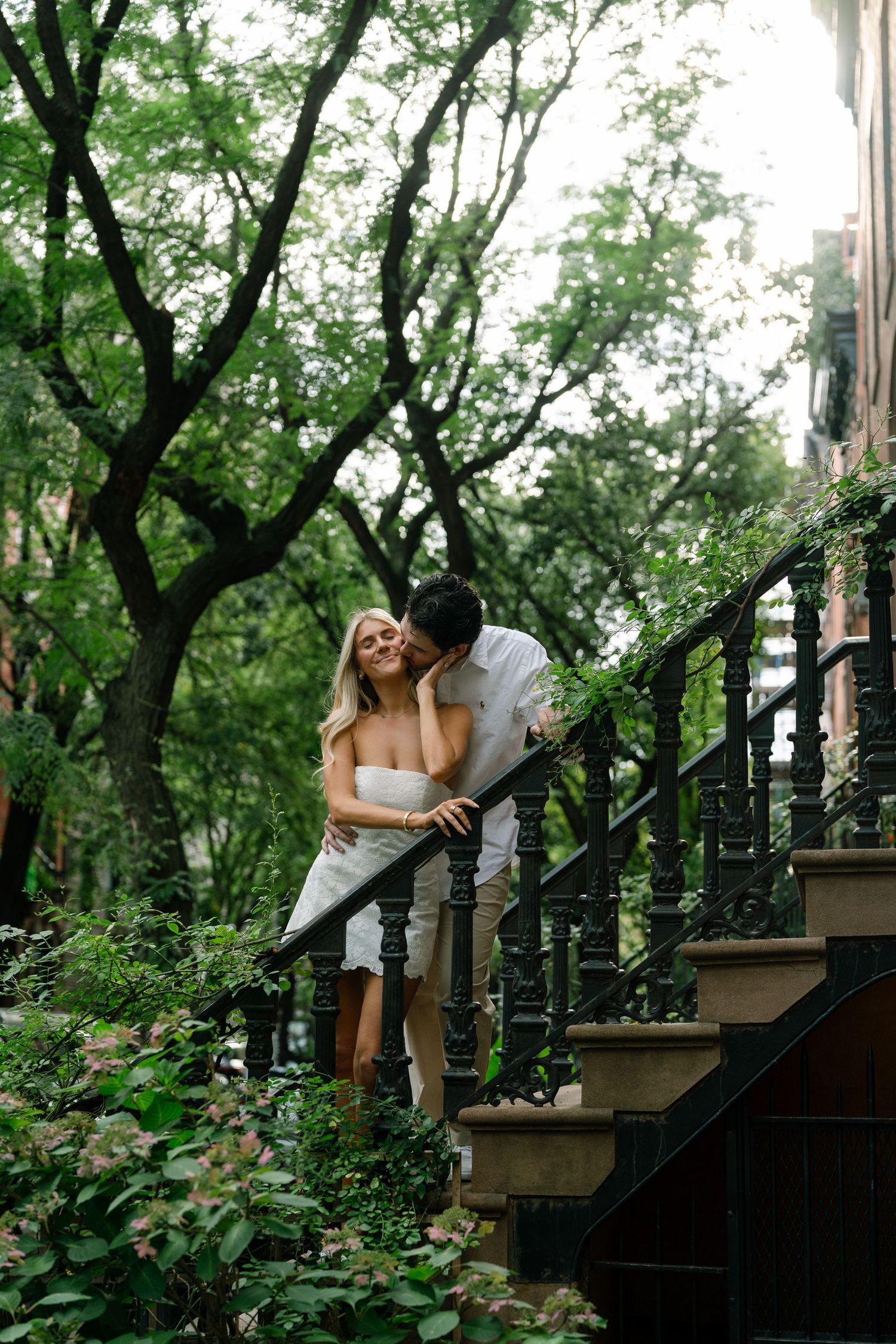 A couple shares a romantic moment on iron staircase steps surrounded by lush green trees in an urban setting.