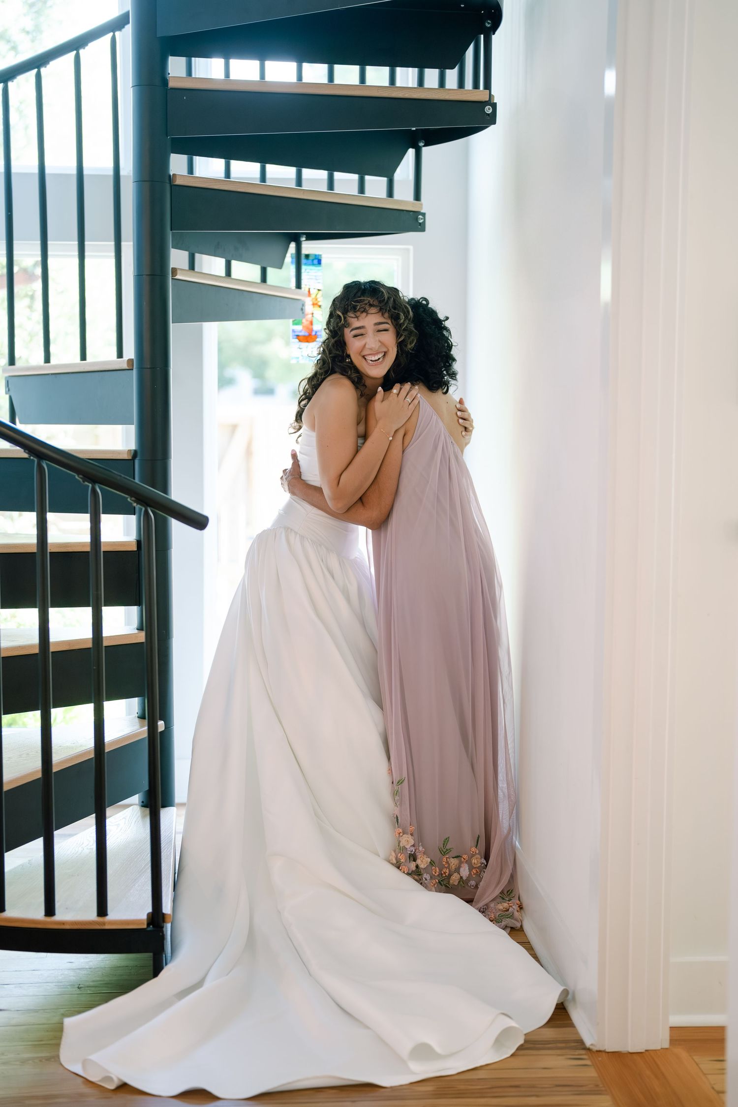 Bridesmaids share emotional hugs with the bride in white dress against bright interior with spiral staircase.