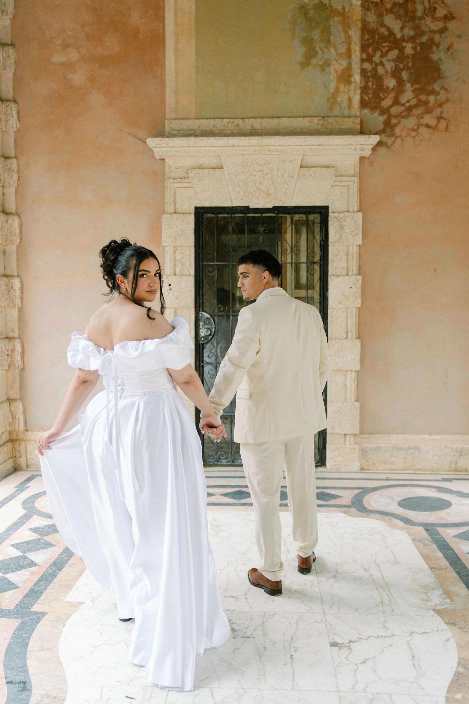 A couple in white wedding attire hold hands while standing on an ornate patio with a peach-colored wall and stone doorway.