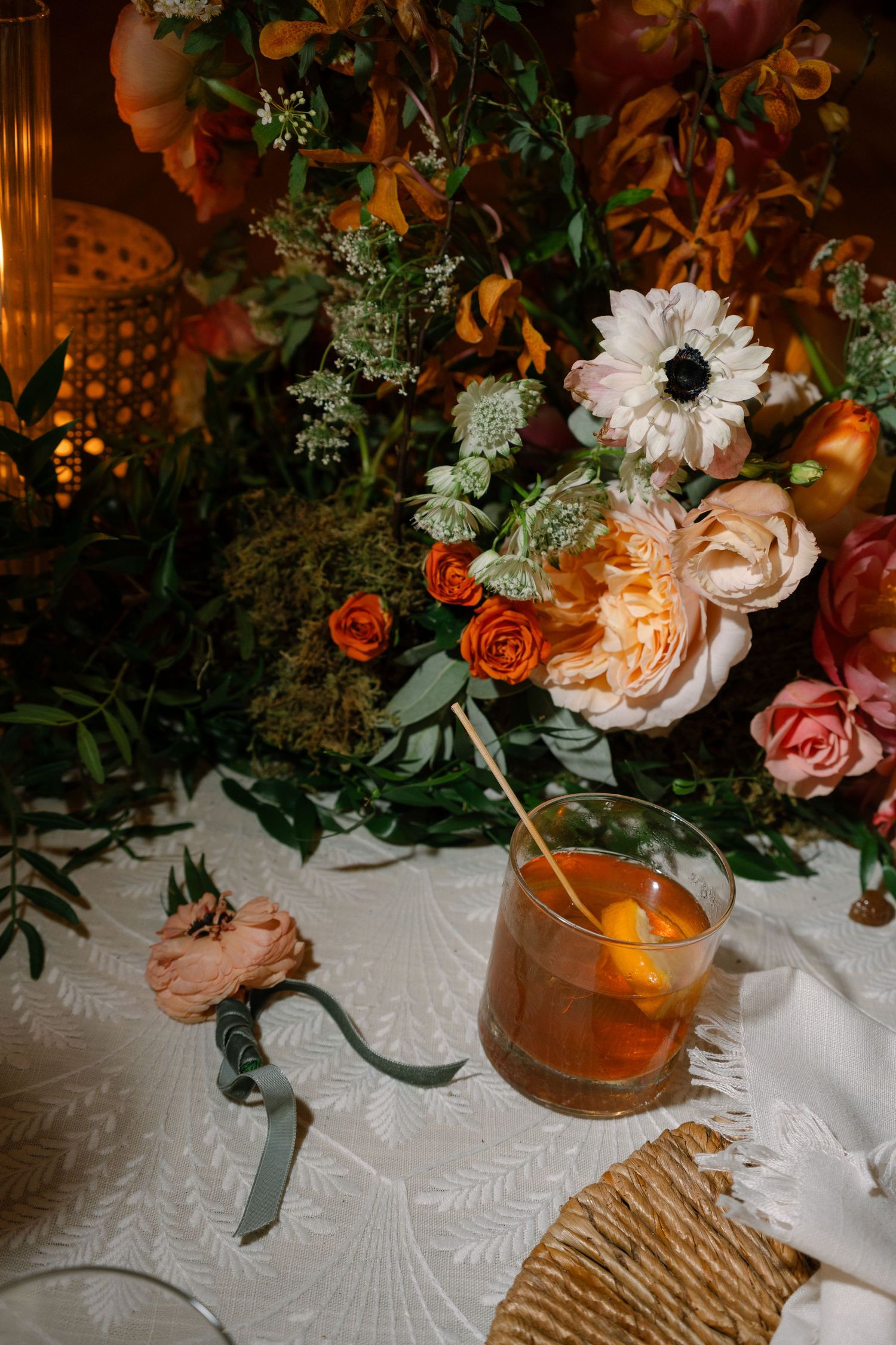 Elegant floral arrangement with roses and anemones next to a craft cocktail on a textured tablecloth.