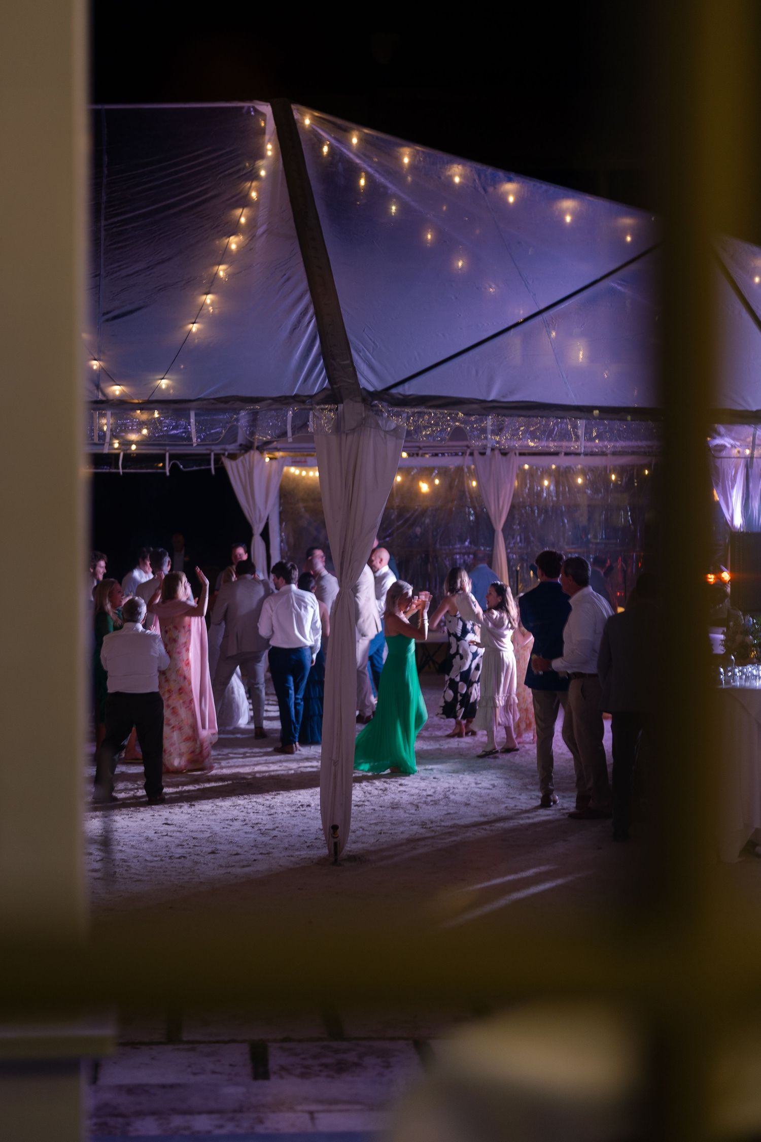 Wedding guests dance under string lights in a clear tent during evening reception.