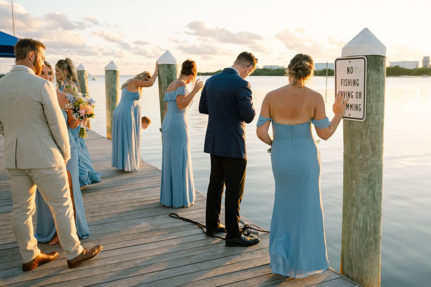 Wedding party in blue dresses standing on a waterfront dock at sunset.