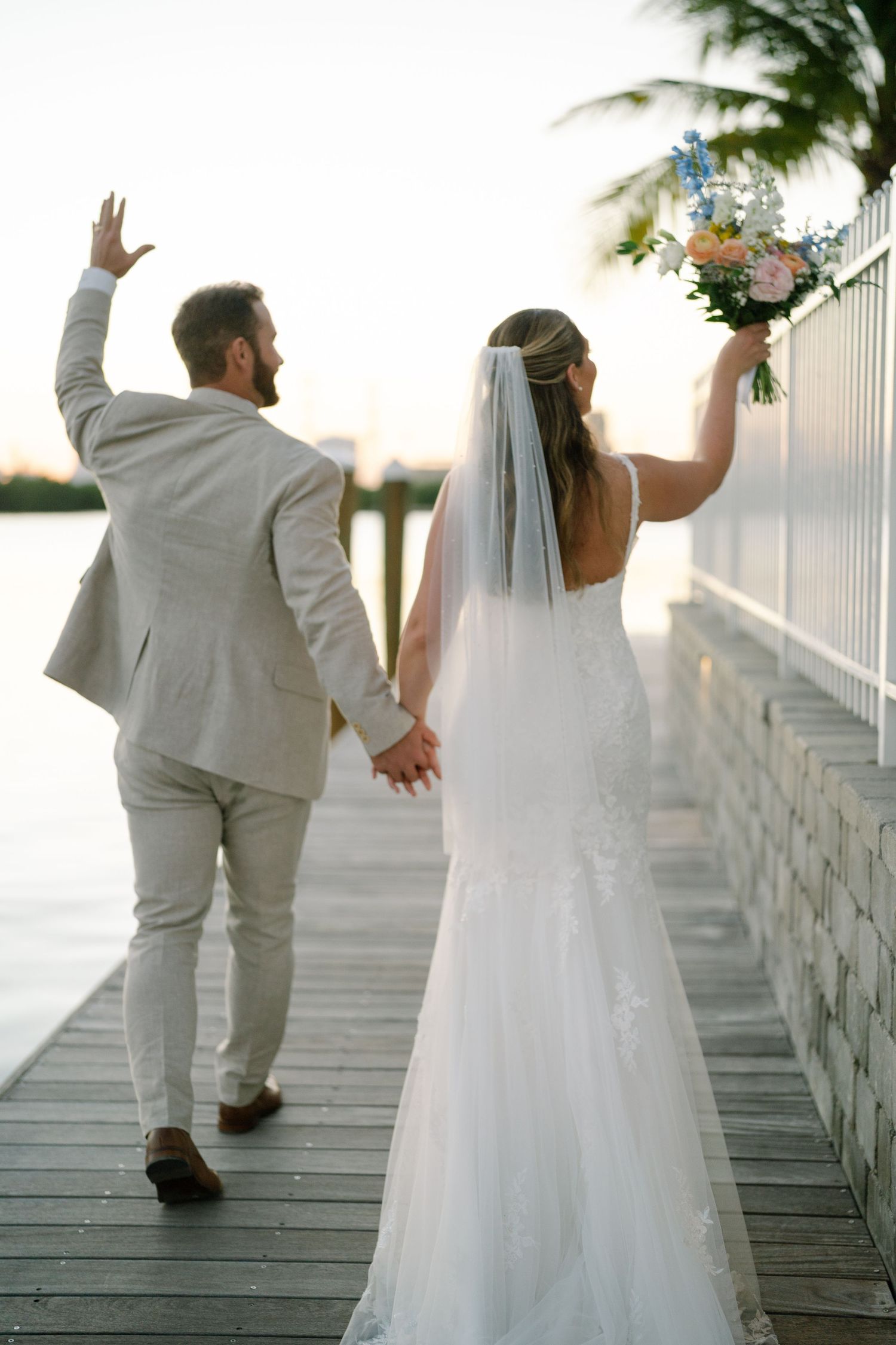 A couple in wedding attire walk along a waterfront boardwalk at sunset holding hands and celebrating.