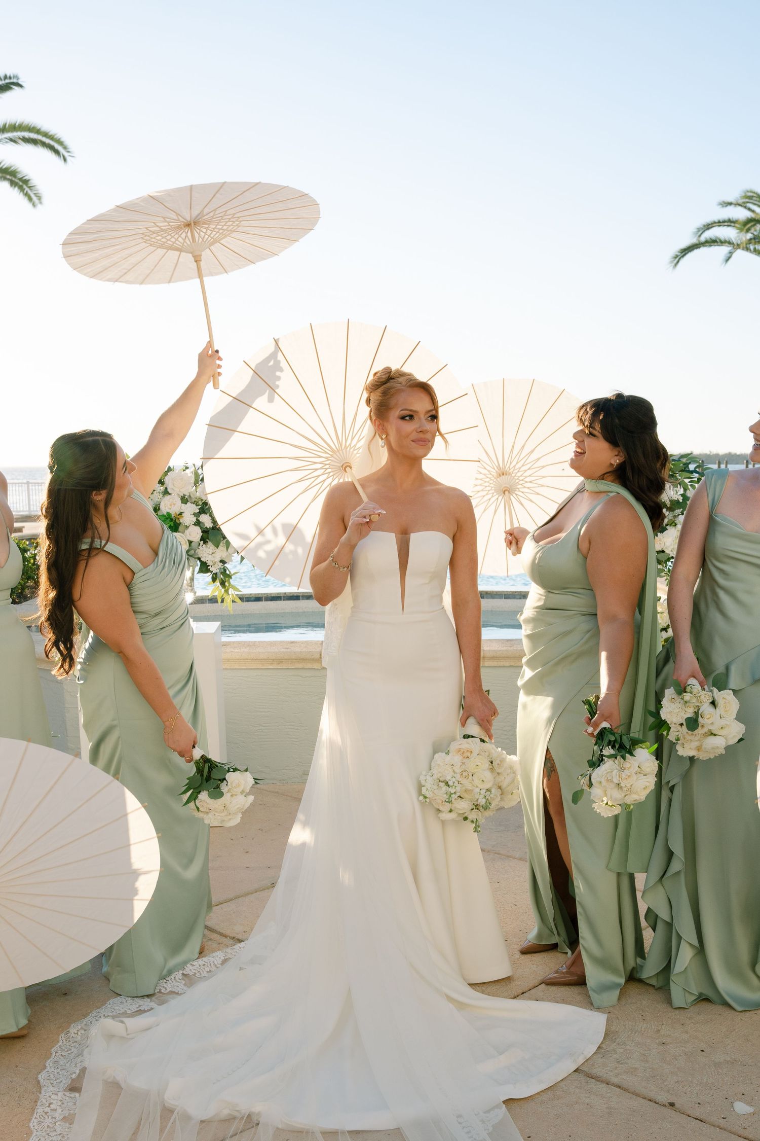 Wedding party in sage green dresses holding white parasols and bouquets during an outdoor celebration.