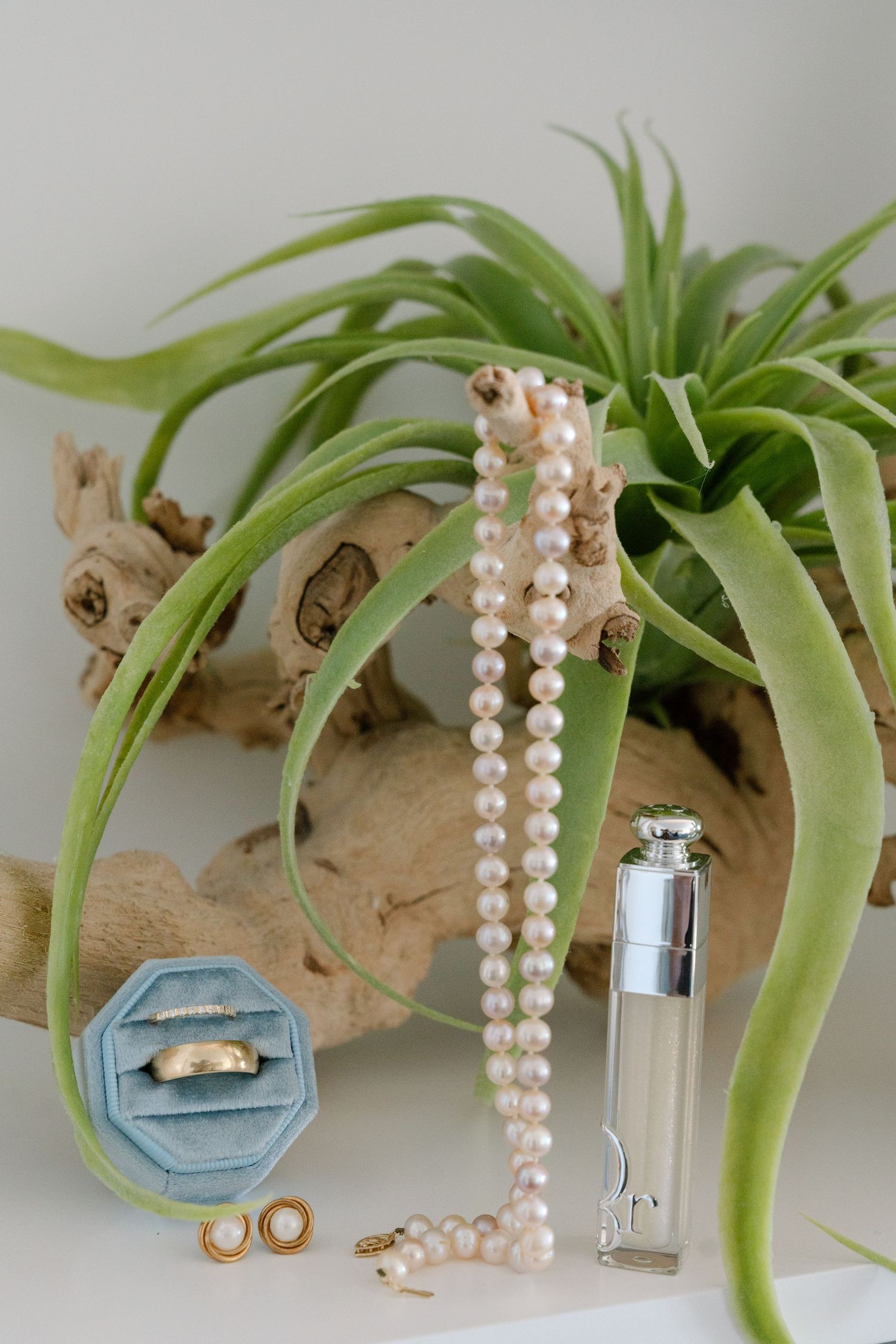 Pearl necklace and engagement ring displayed on blue velvet ring box with decorative green plant branches.