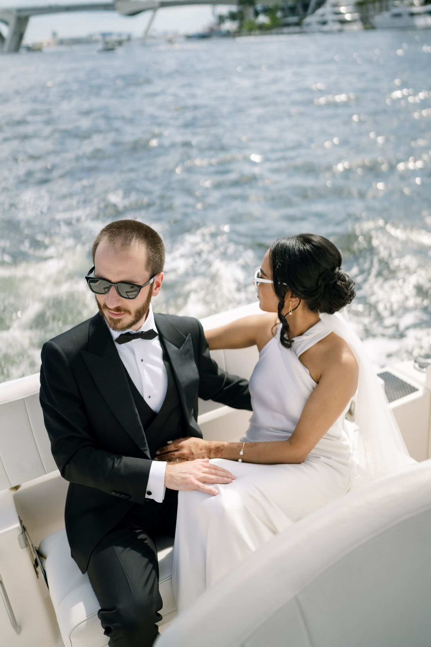 A couple in formal wedding attire share a romantic moment on a boat with sparkling water in the background.