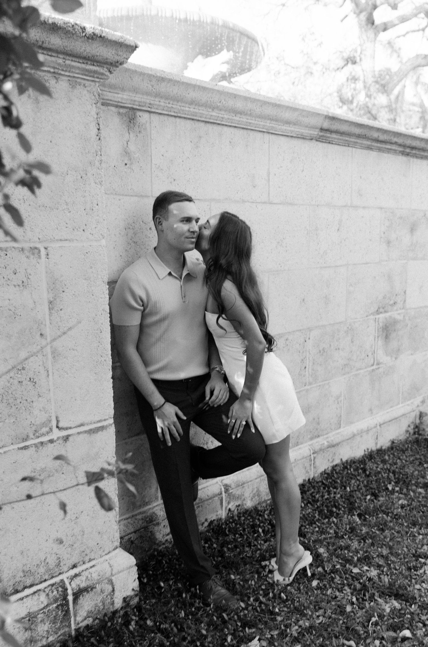A romantic black and white photograph of a couple sharing an intimate moment against a stone wall outdoors.