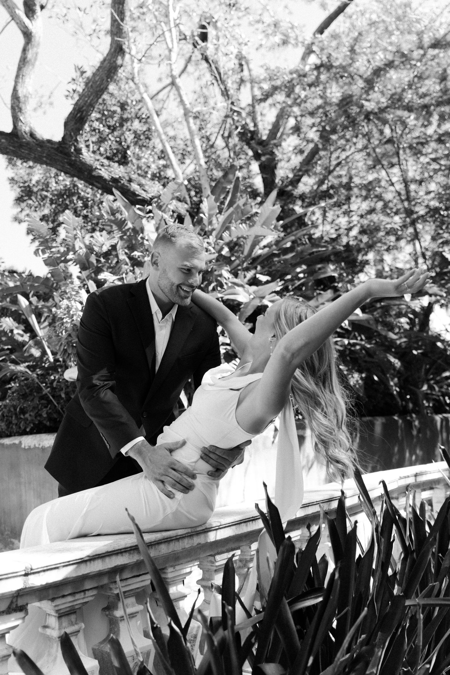 A black and white romantic photo of a couple dancing outdoors near a stone railing surrounded by lush garden foliage.