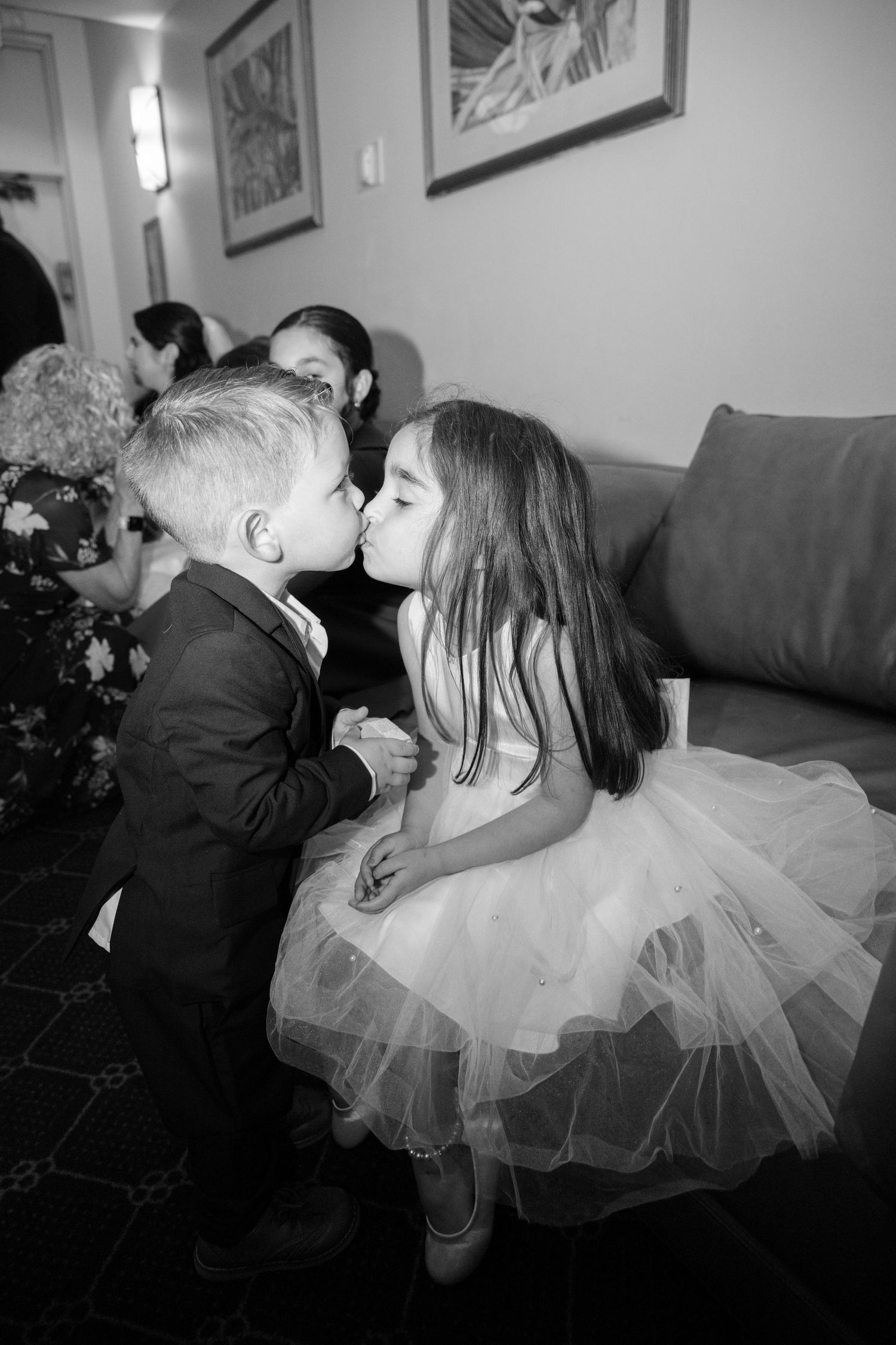 A young boy in a suit and girl in a tulle dress share an innocent kiss at what appears to be a formal event.