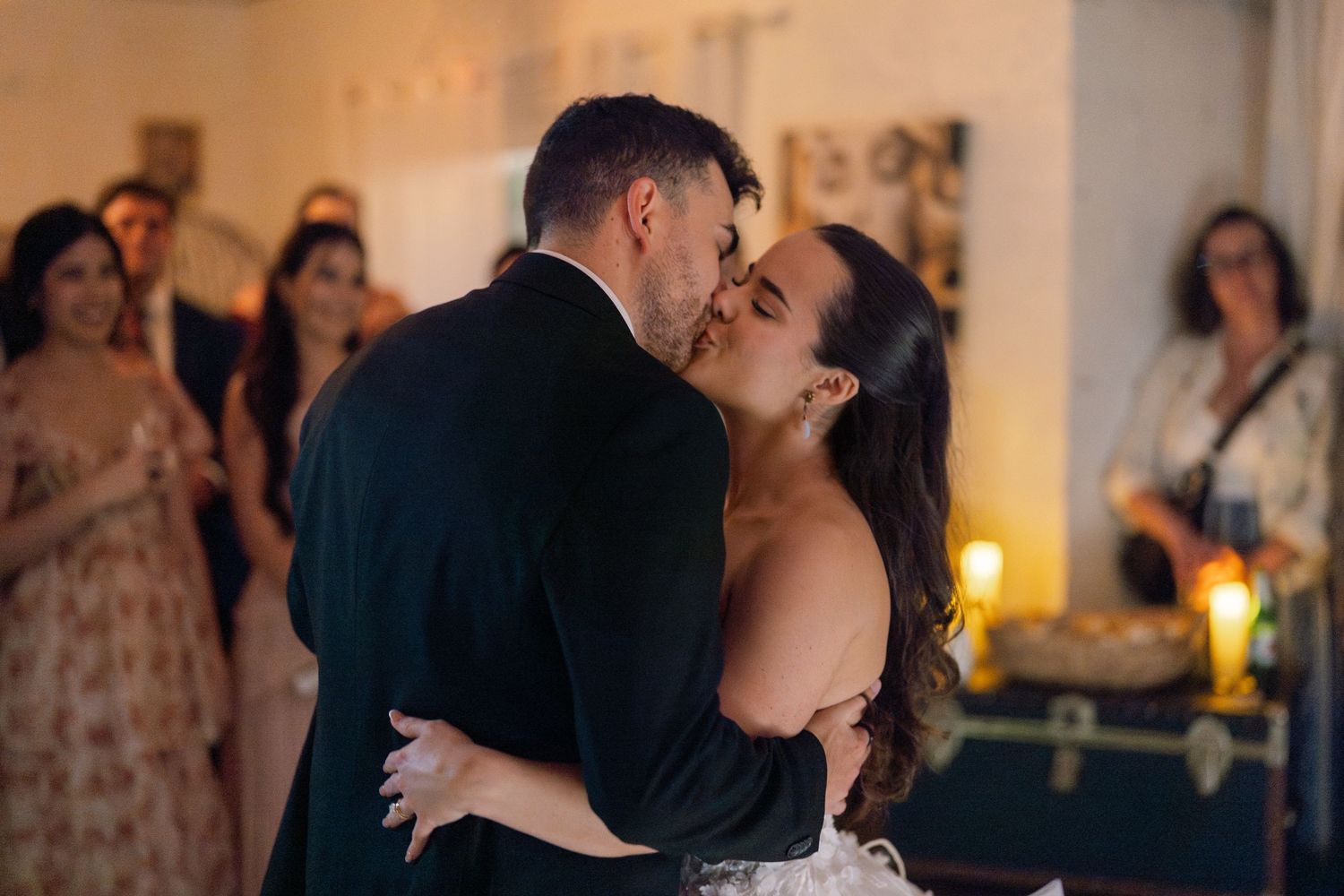 A couple shares a romantic first dance kiss at their wedding reception, surrounded by guests in a warmly lit venue.