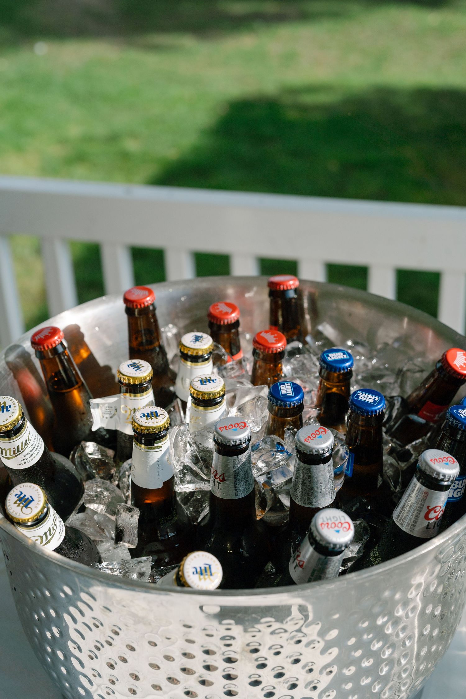 A metal bucket filled with assorted beer bottles and ice, placed on a white porch railing with green grass in background.