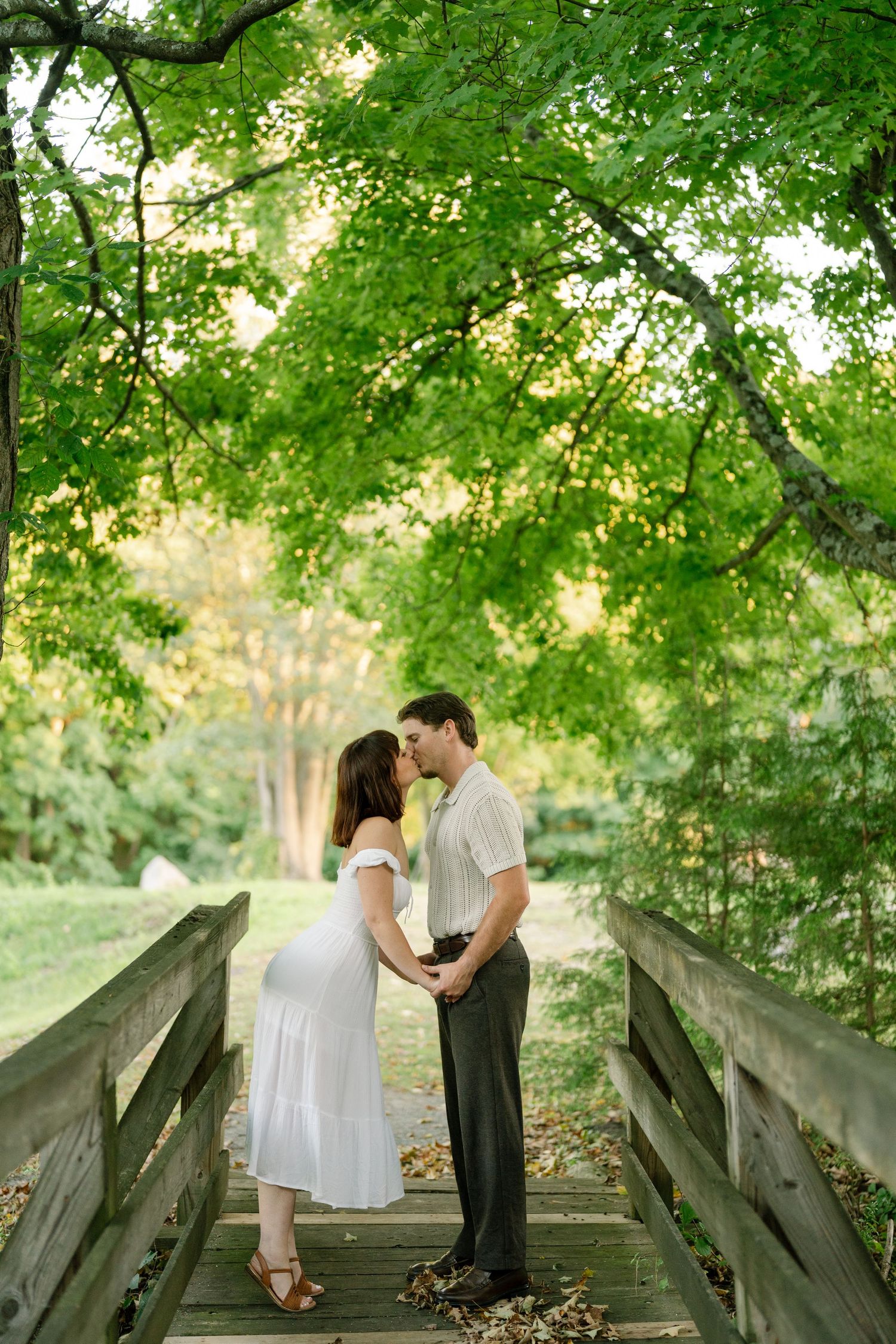 Pregnant couple sharing a kiss on a wooden bridge surrounded by lush green trees in a park setting.