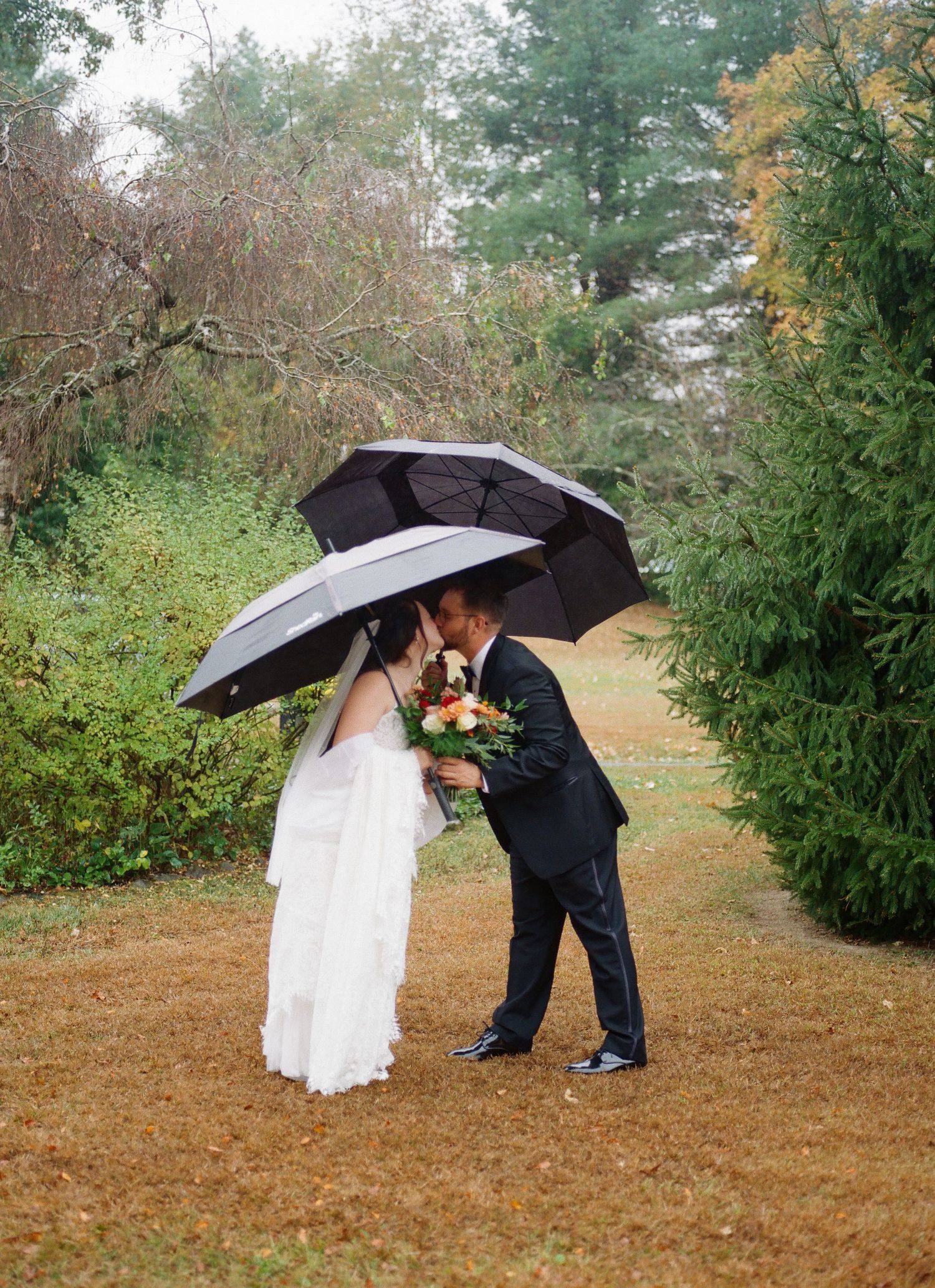 A couple shares a romantic moment under a black umbrella on a rainy autumn day in a wooded outdoor setting.