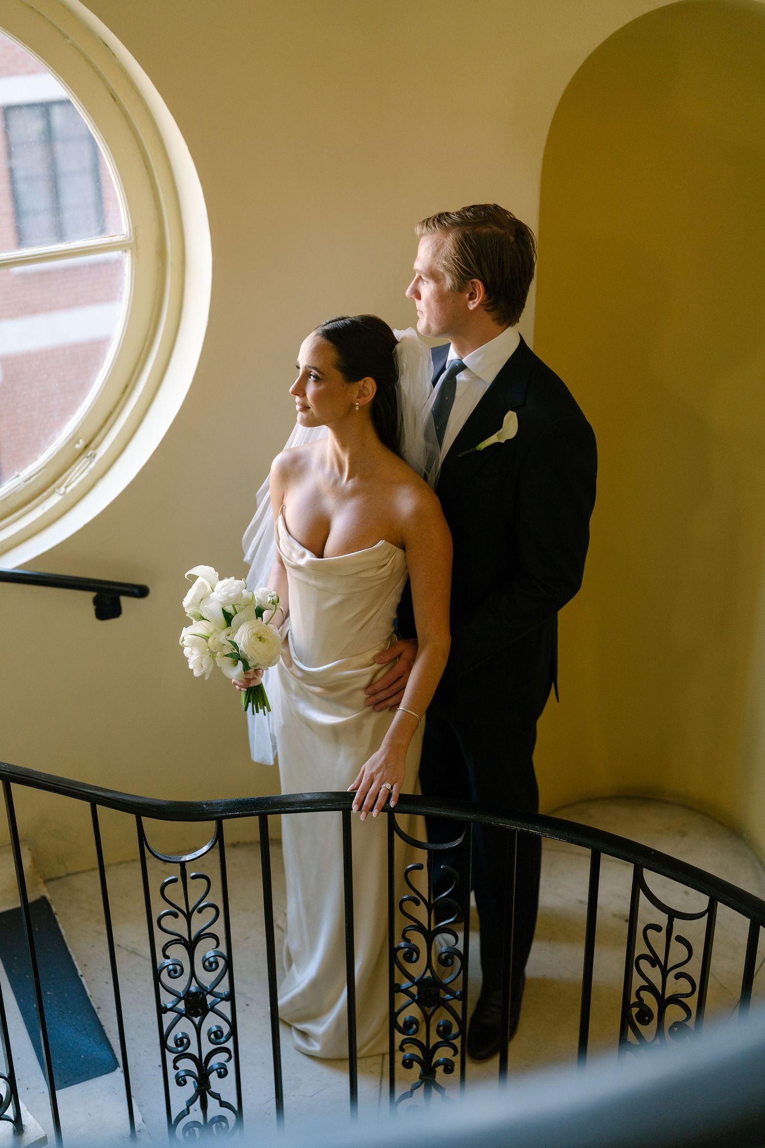 Bride and groom pose elegantly on a curved staircase, she holds white roses, both gazing toward a round window.