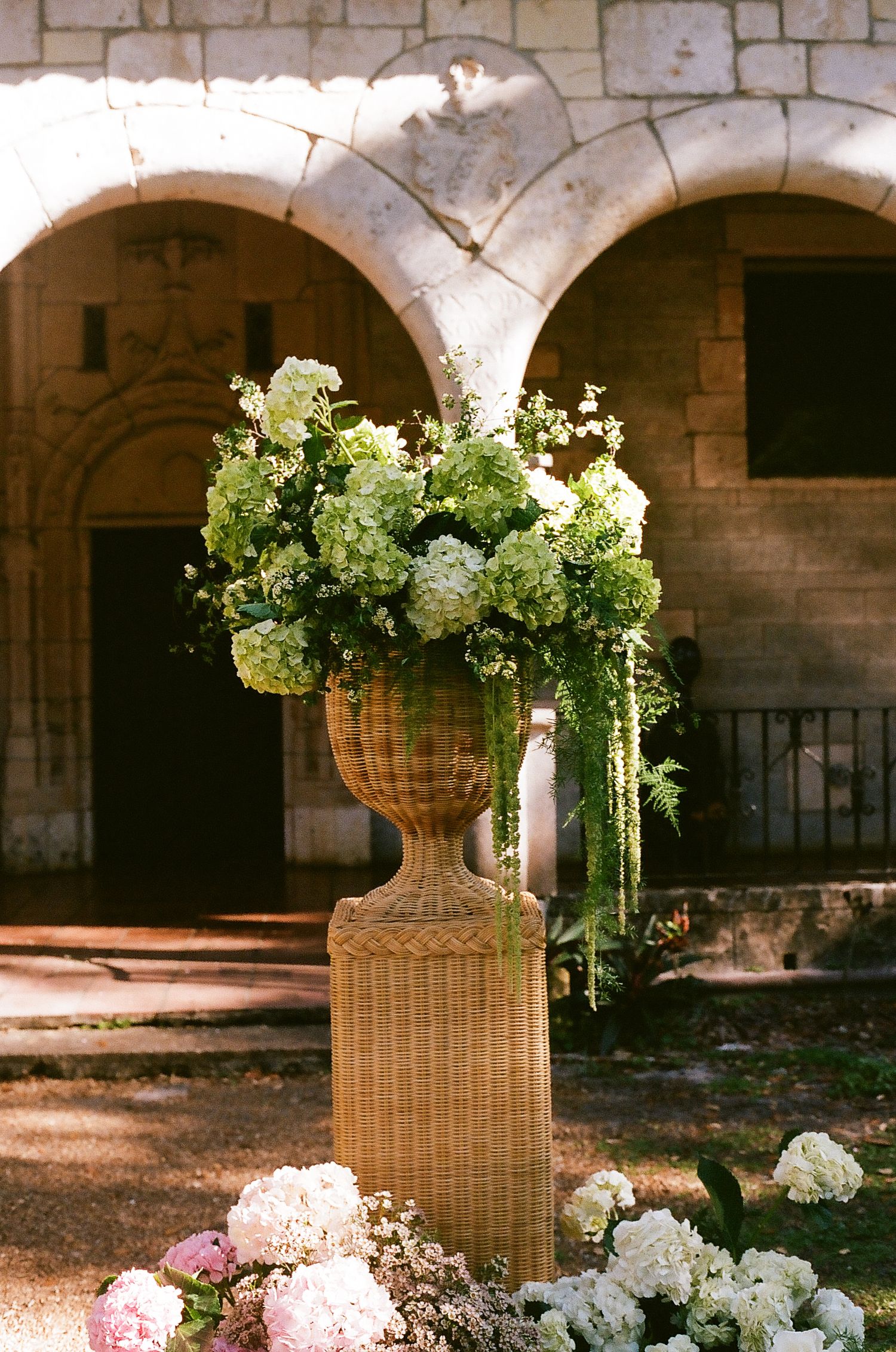 Elegant wicker urn overflowing with green and white hydrangeas on a pedestal outside a stone archway venue.