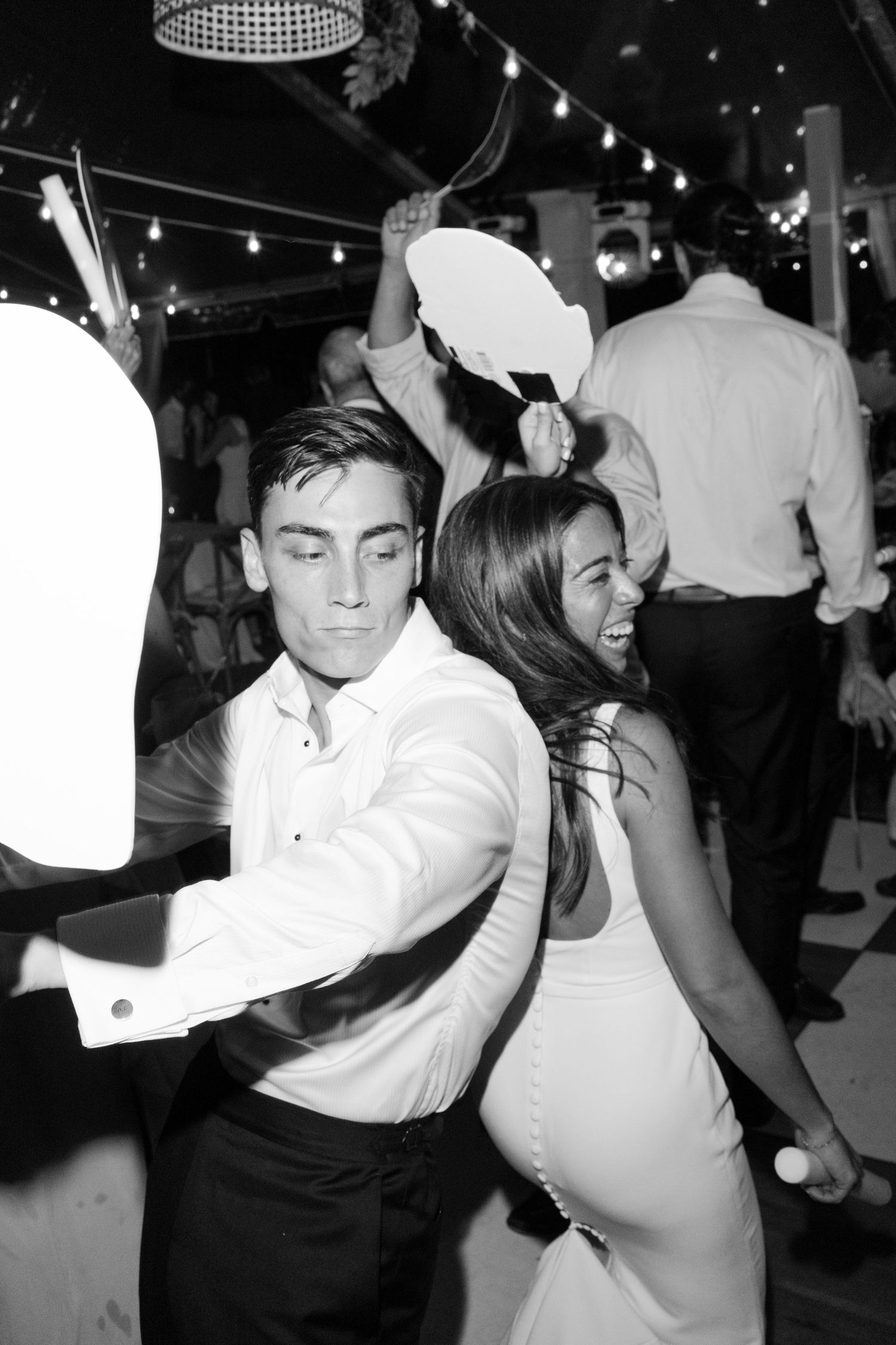 Black and white photo of a couple dancing energetically at a wedding reception with string lights in the background.