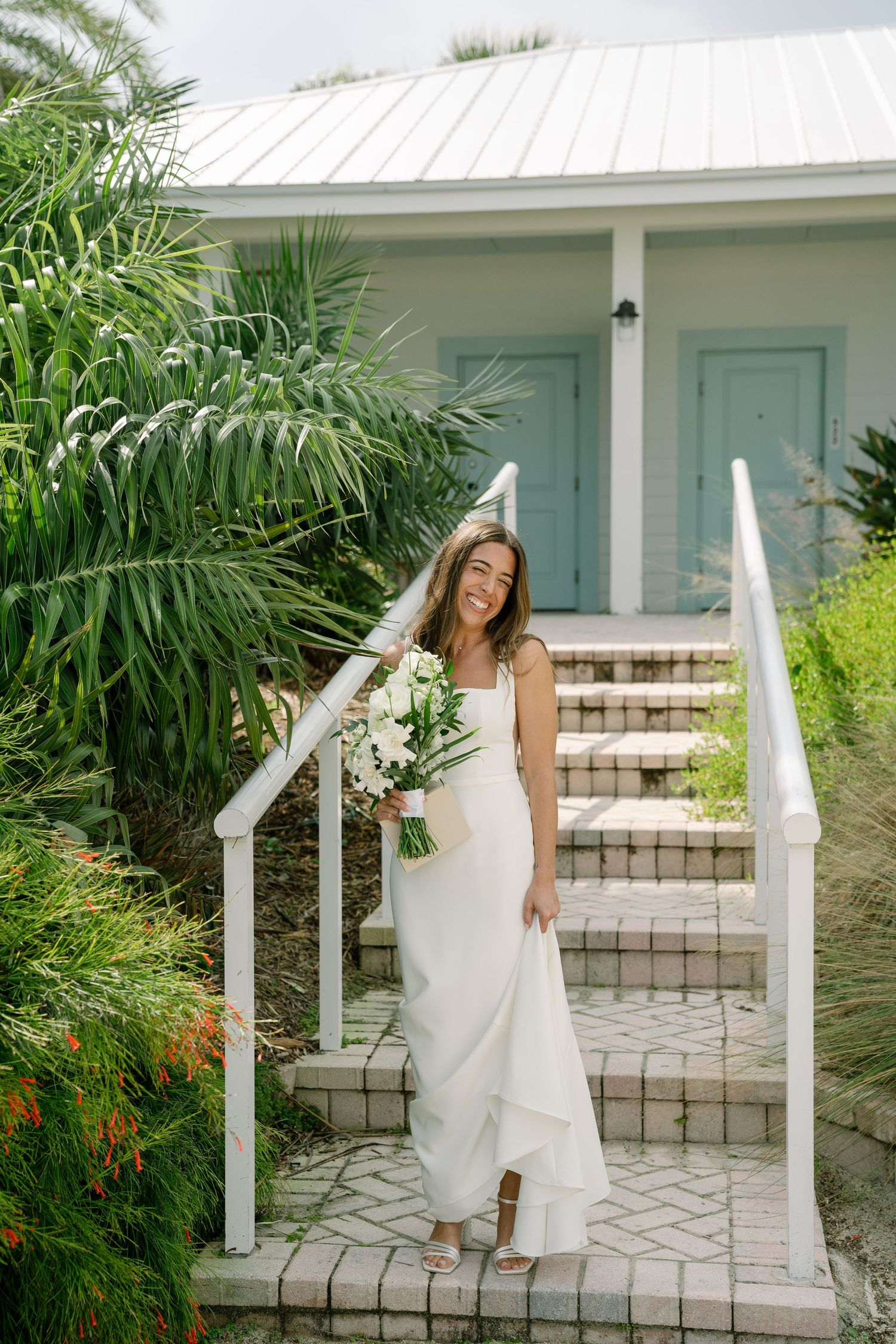 Smiling bride in white gown holding bouquet on brick steps with tropical greenery and blue cottage.