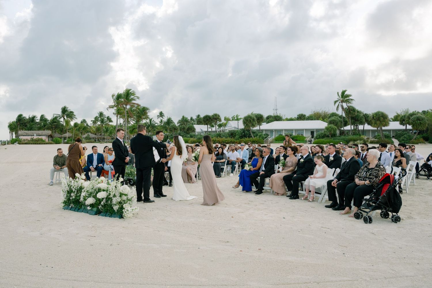 Outdoor beach wedding ceremony with guests seated on white sand facing couple at floral altar.