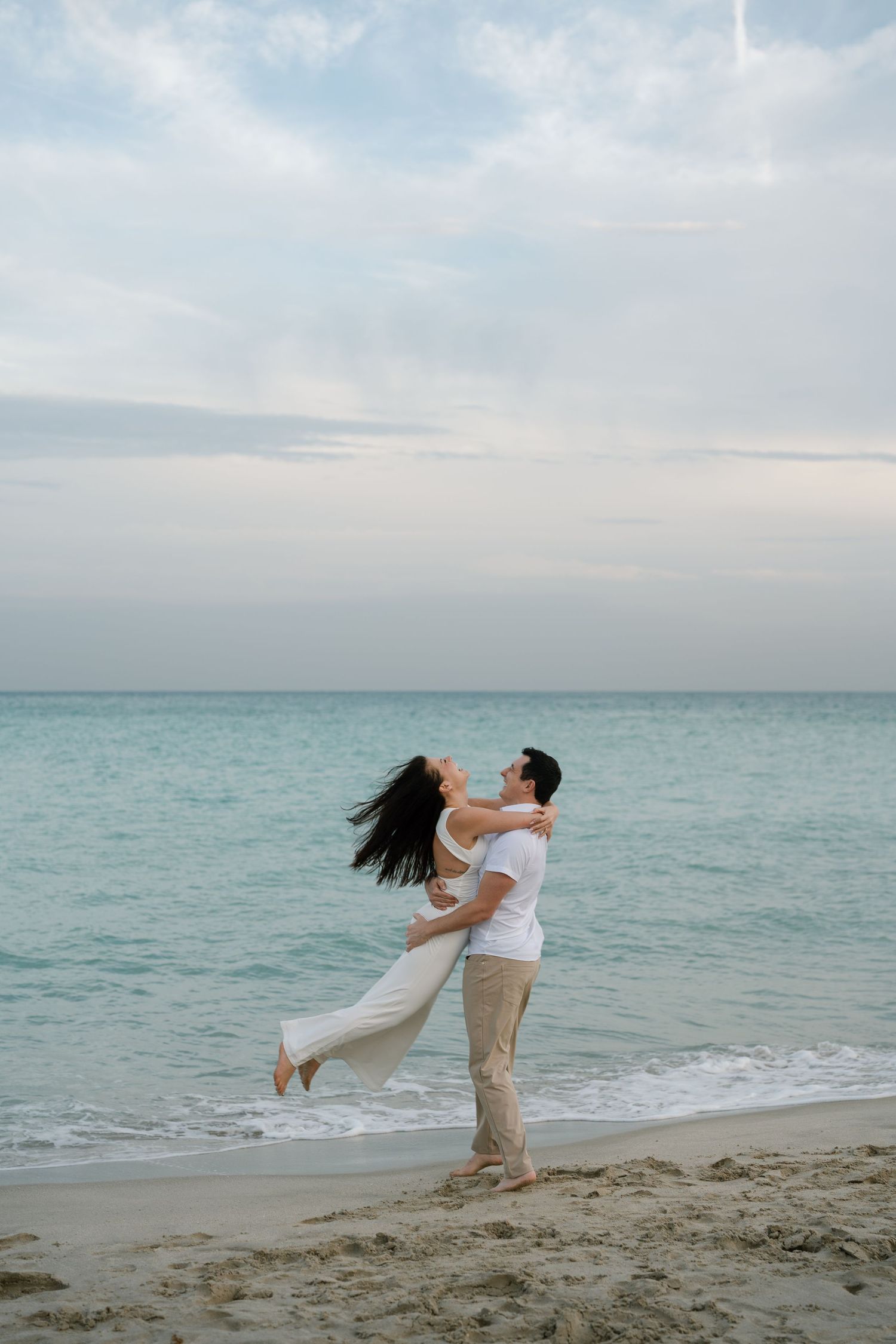 Romantic couple in white clothing dancing playfully on a beach with turquoise ocean waters.