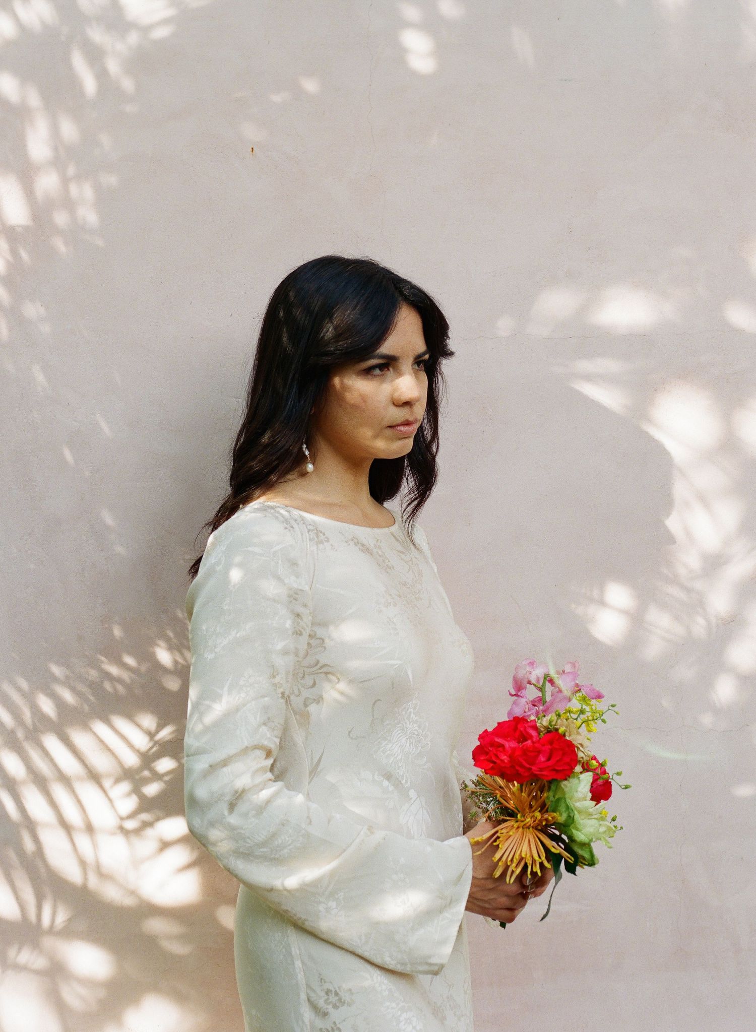 Figure in flowing white dress holding colorful bouquet against wall with dappled sunlight.