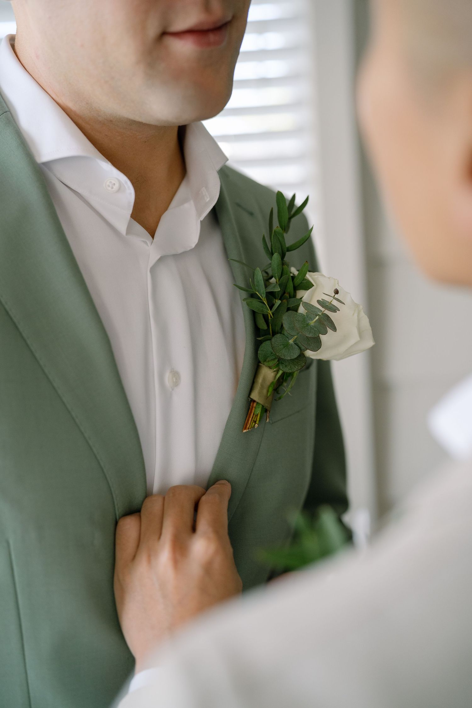 A groom wearing a sage green suit jacket with a white boutonniere adjusts his lapel in an elegant wedding detail shot.