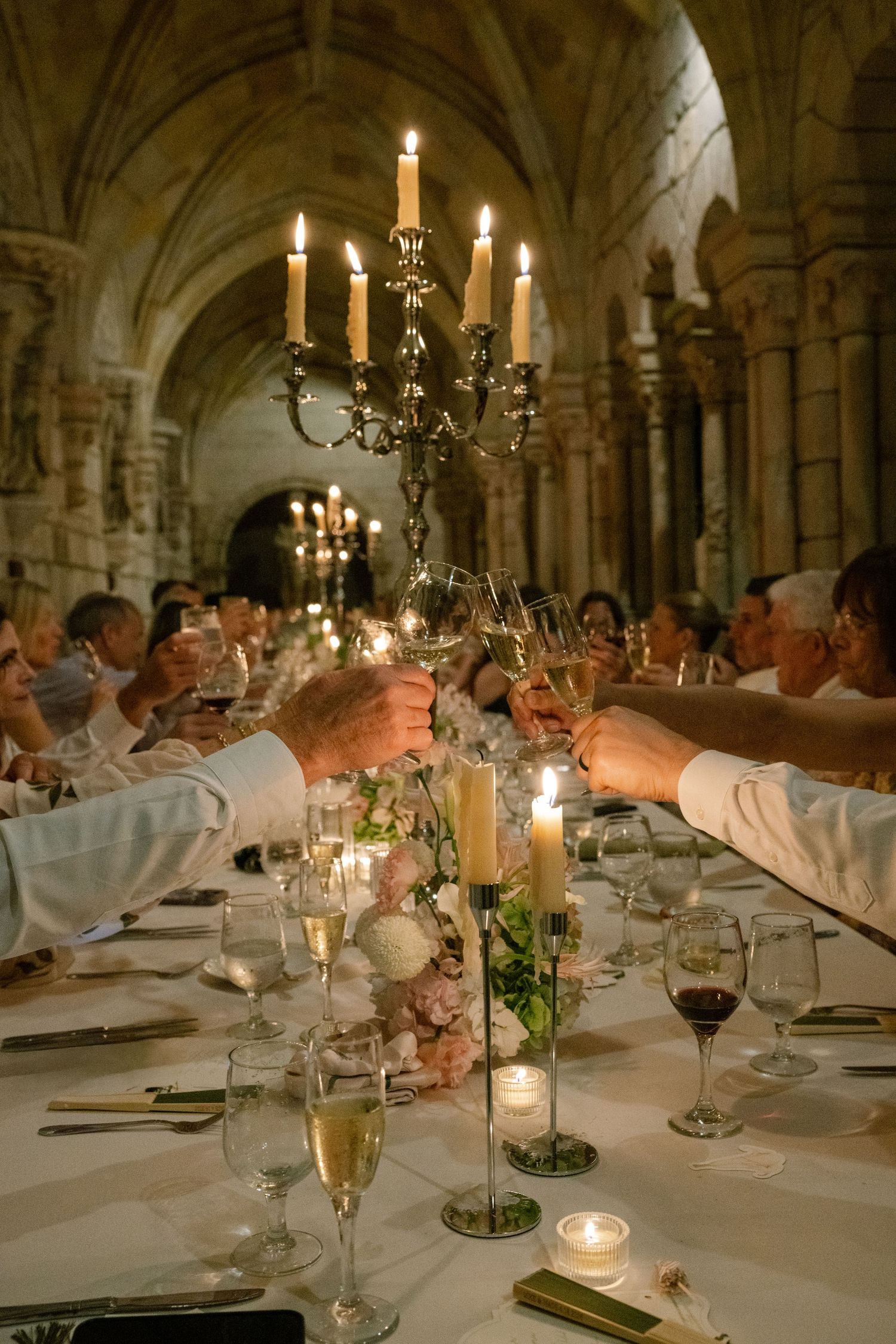 Wedding guests raise champagne glasses in candlelit toast at long table in stunning stone cathedral.