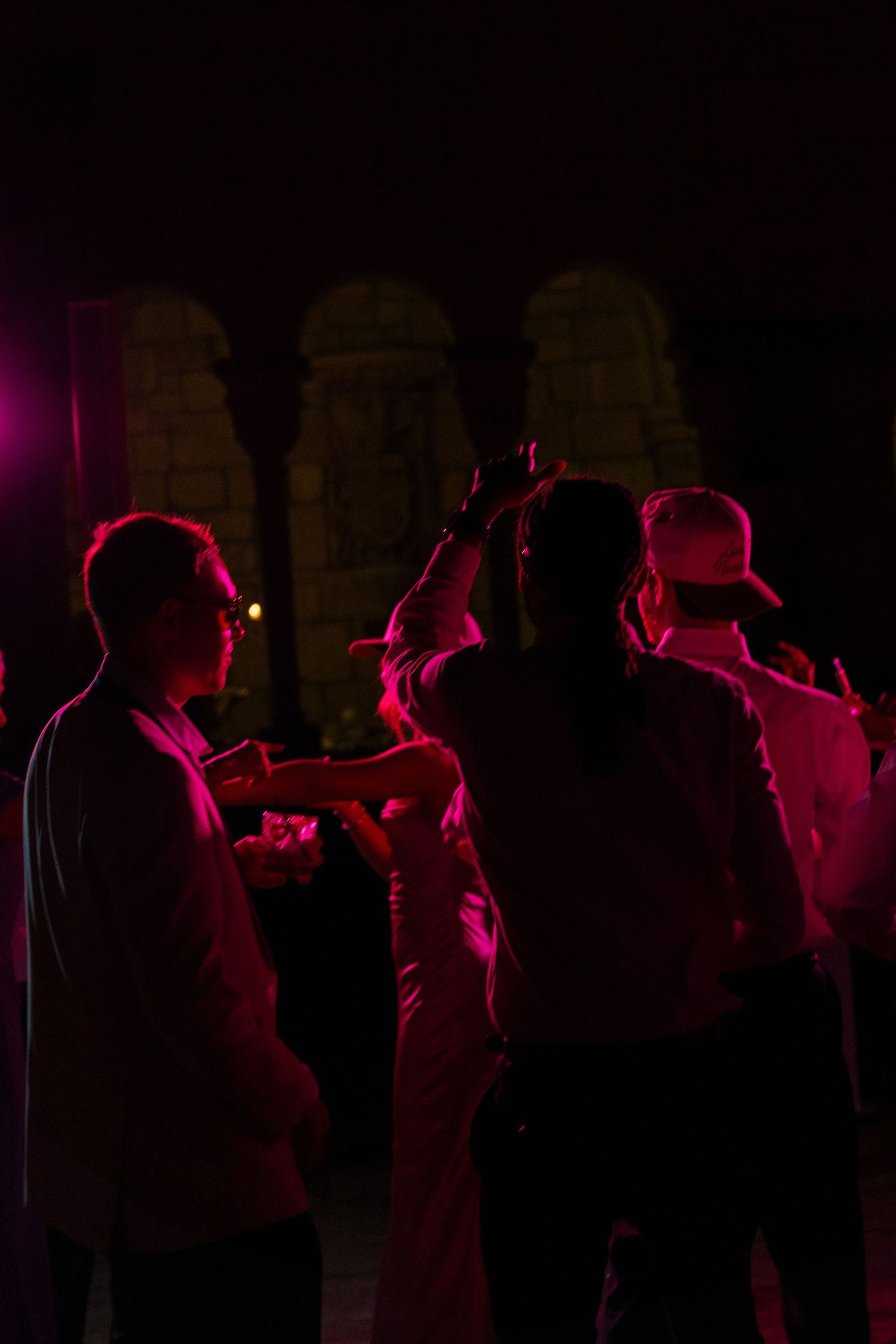 Wedding guests dancing under pink and red lights at nighttime outdoor reception near stone columns.