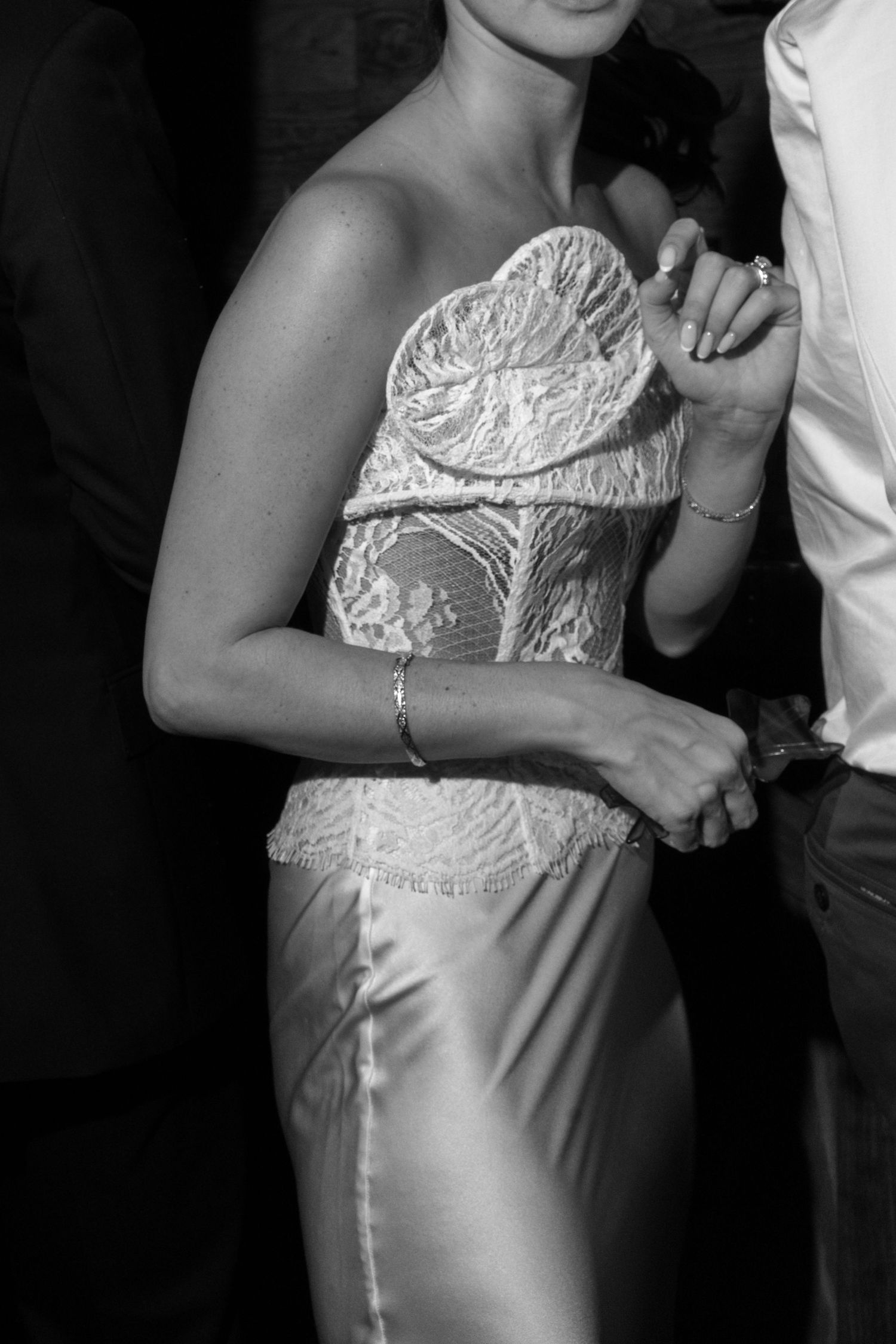 Black and white close-up of bride in strapless lace corset wedding gown holding hands with groom during reception dance.