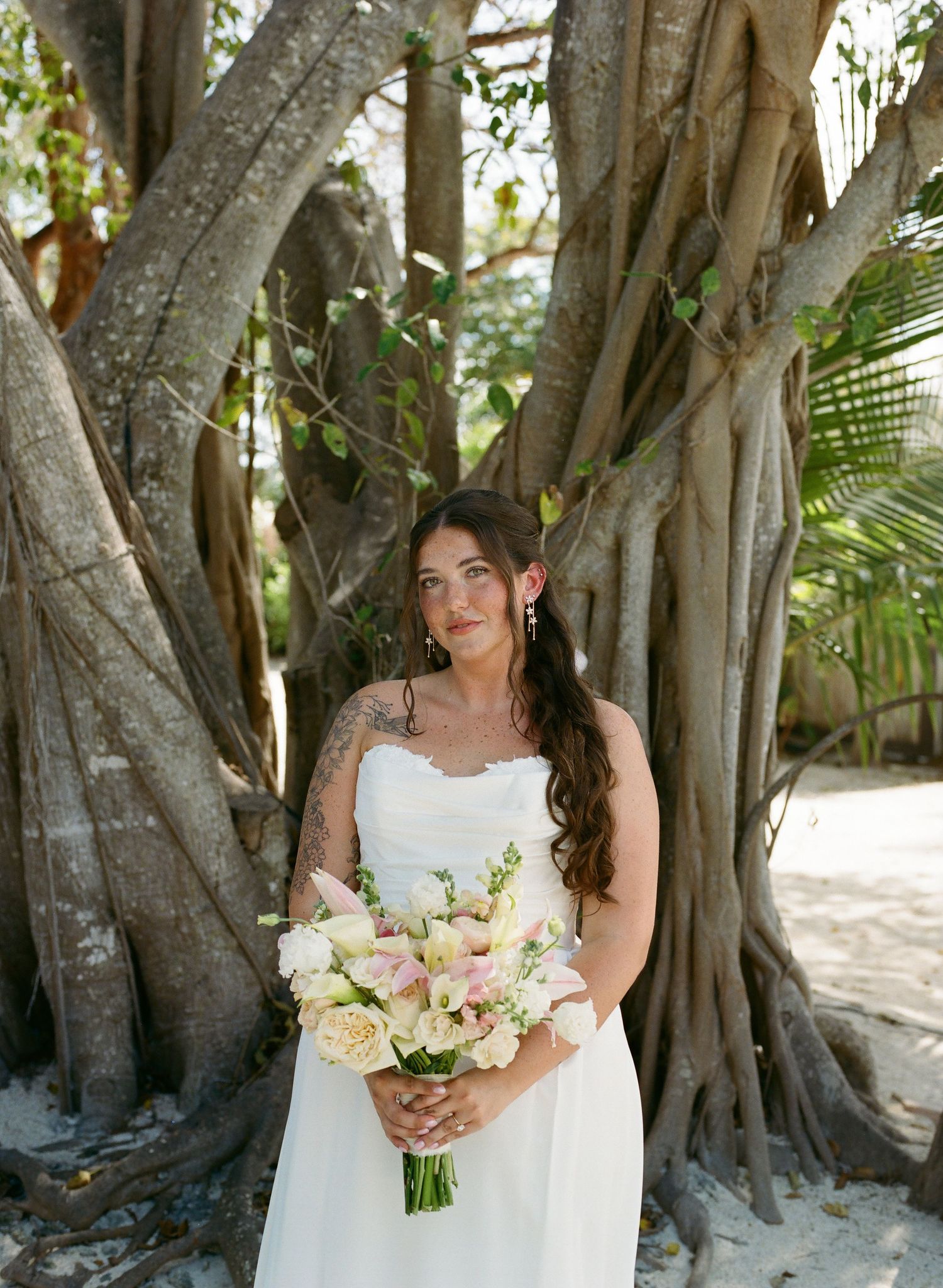 Bride in white strapless gown holding pink bouquet poses before large banyan tree roots outdoors.