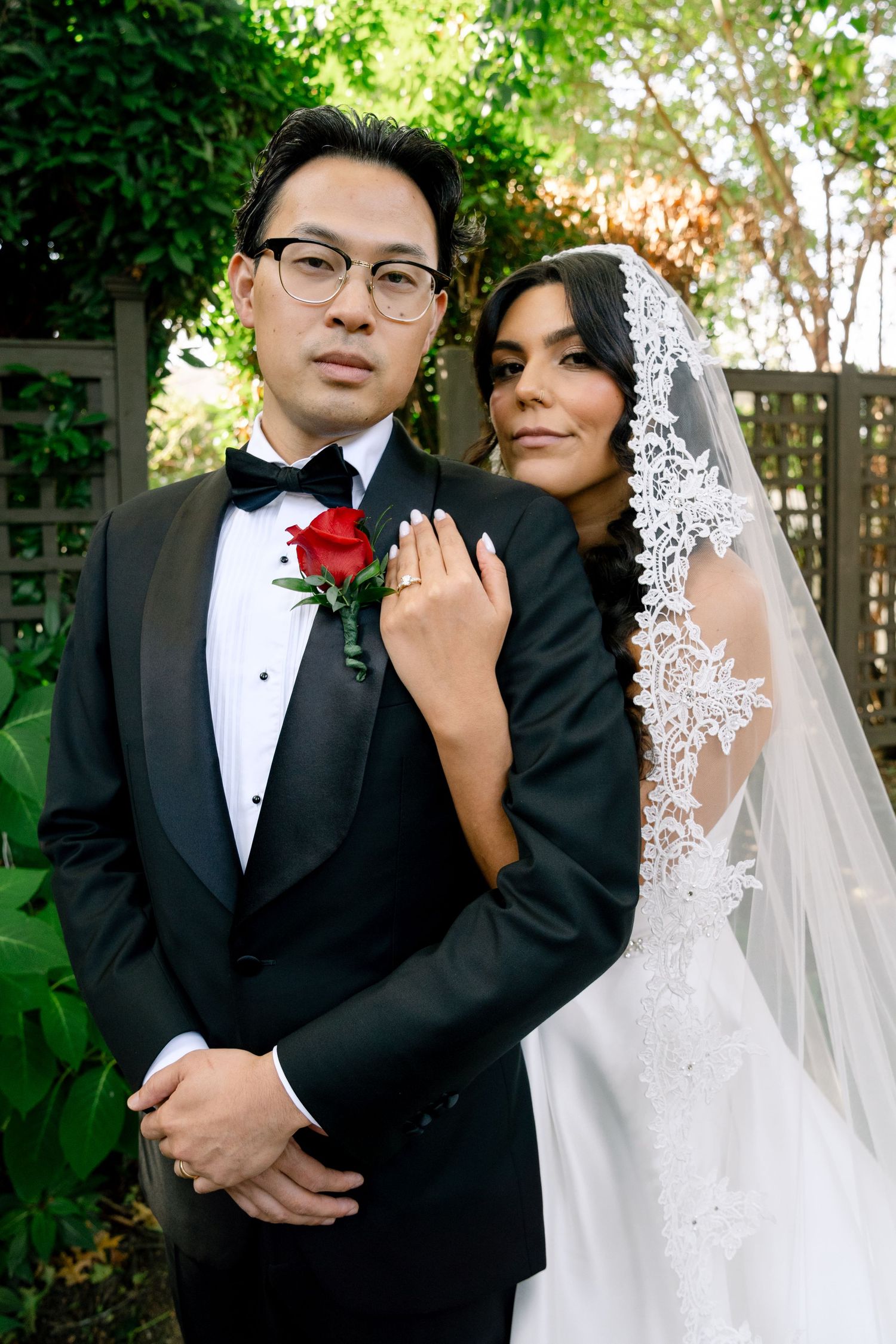 A couple in formal wedding attire poses in a garden setting with greenery and sunlight.