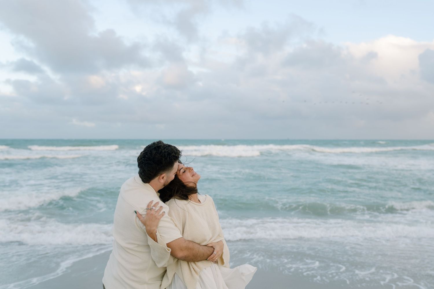 A couple in light colored attire embraces on a cloudy beach with waves crashing in the background.