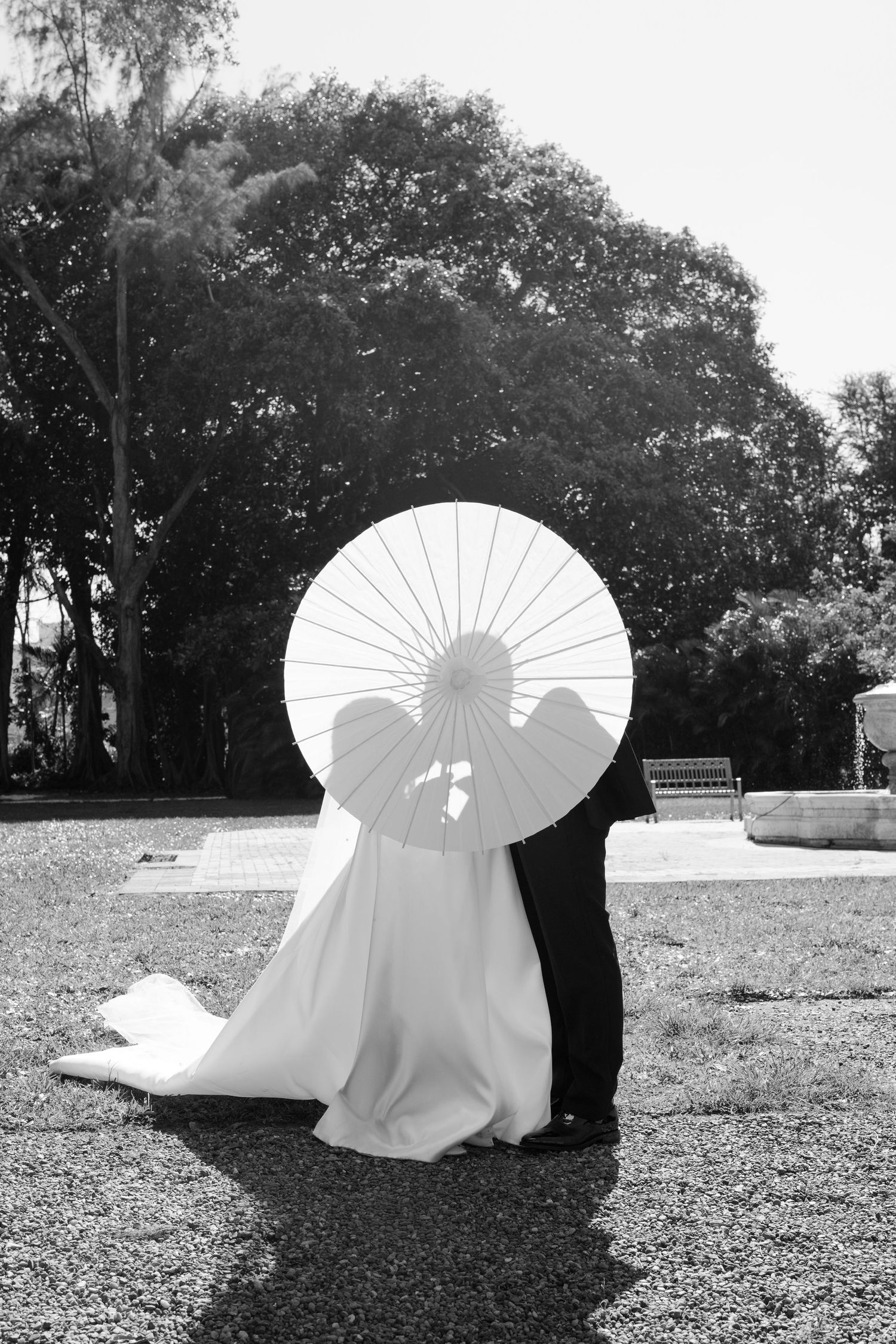 Black and white photo of a bride and groom kissing behind a white parasol umbrella outdoors.