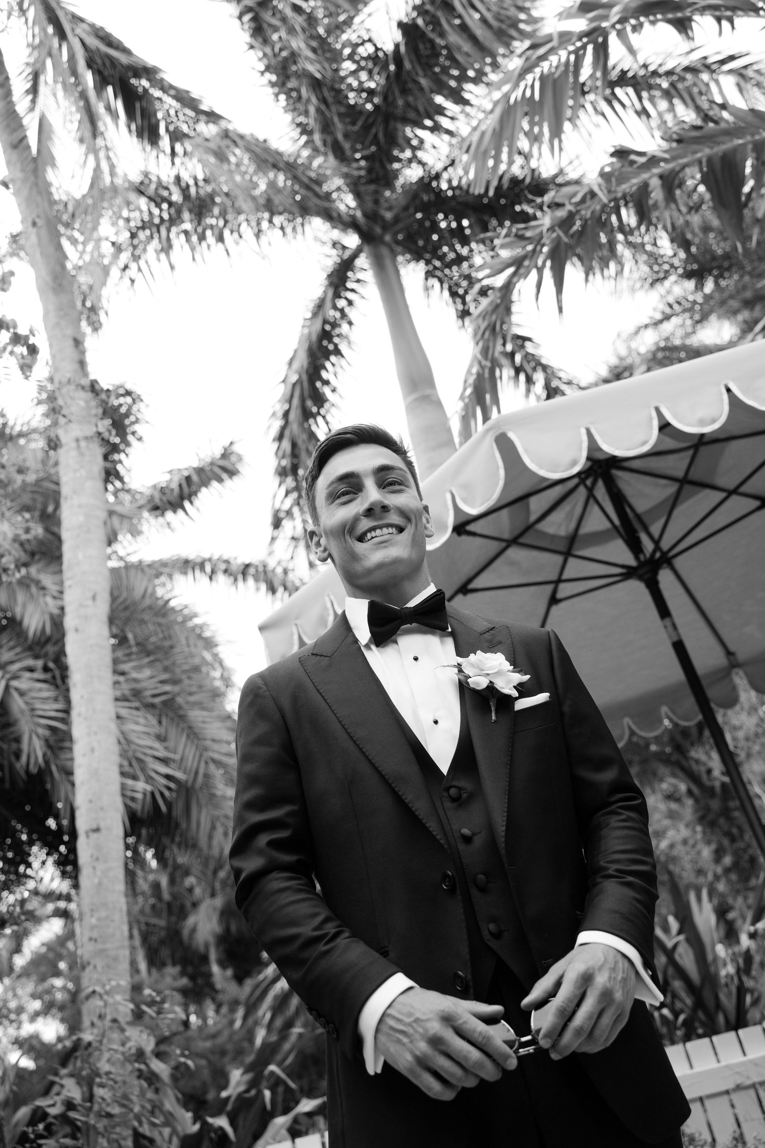 Smiling groom in black tuxedo with bow tie and boutonniere beneath tropical palm trees, black and white.